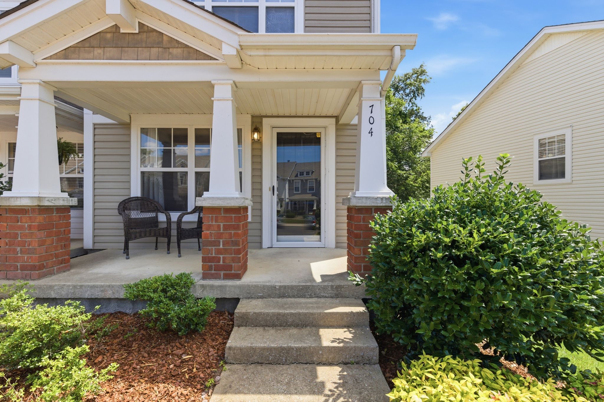 Front porch of a beige house with white trim, brick columns, and glass front door, overlooking a small garden path.