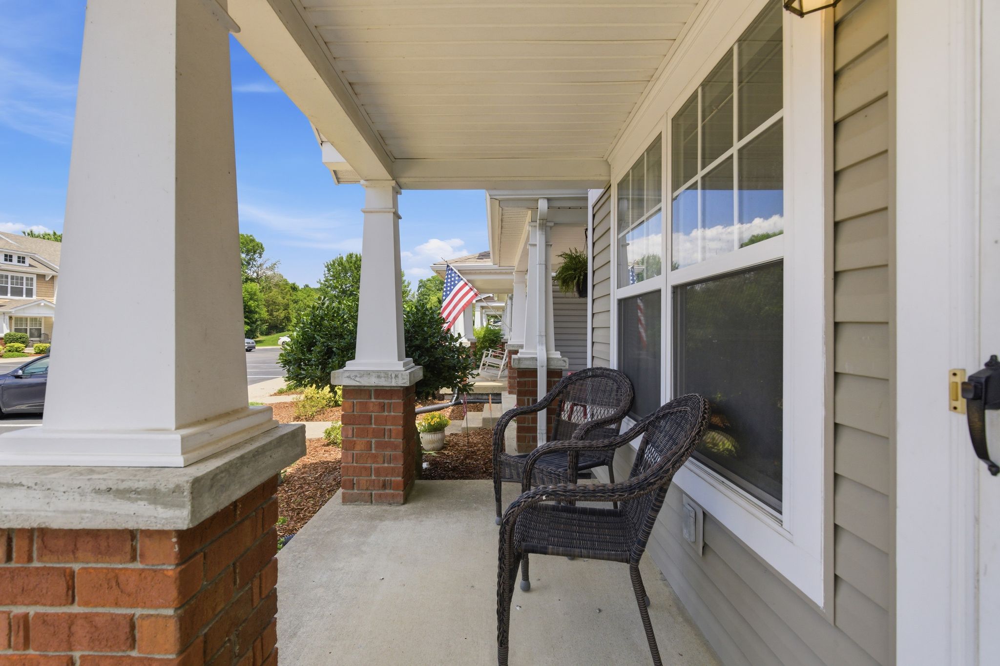 Front porch with white columns, wicker chairs, and an American flag by a large window against a blue sky.