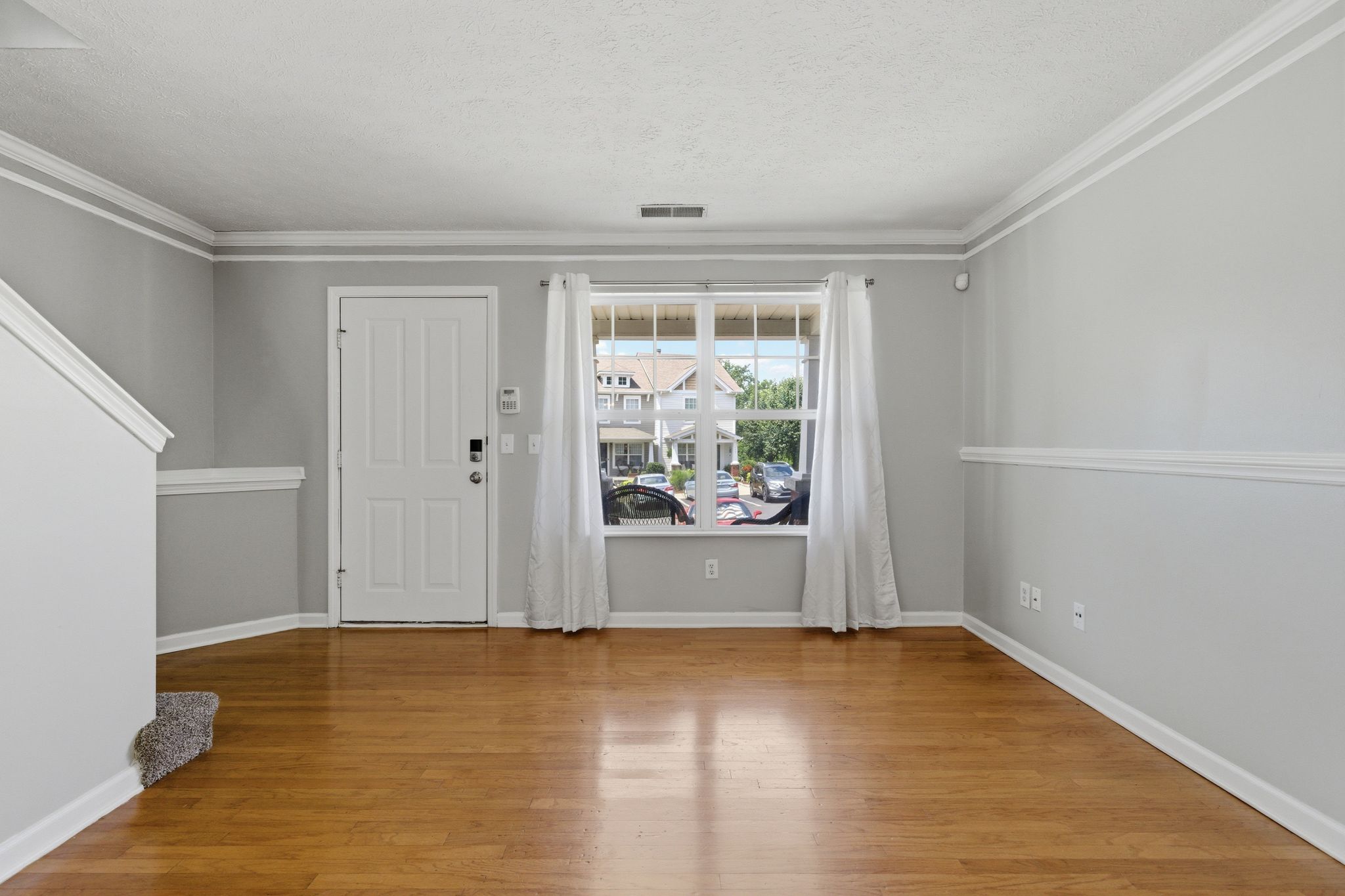 Interior living room with light gray walls, white crown molding, front door to the left, and a large window with white curtains showing a suburban street outside.