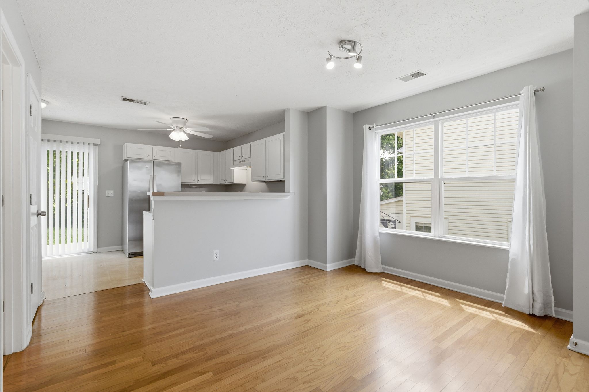 Bright open-concept kitchen and living area with gray walls, white cabinets, stainless fridge, and large window with white curtains. Hardwood floor throughout.