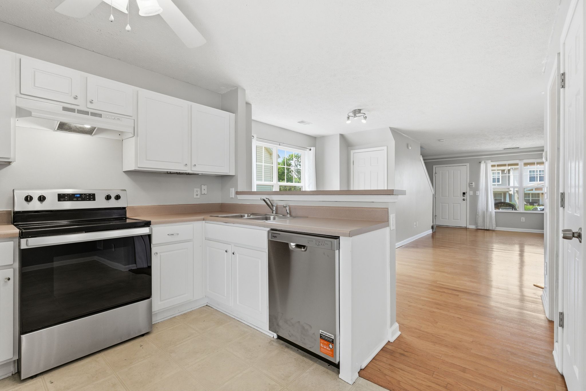 Bright kitchen with white cabinets and stainless steel stove and dishwasher; breakfast bar overlooking living area with hardwood floors and large window light.