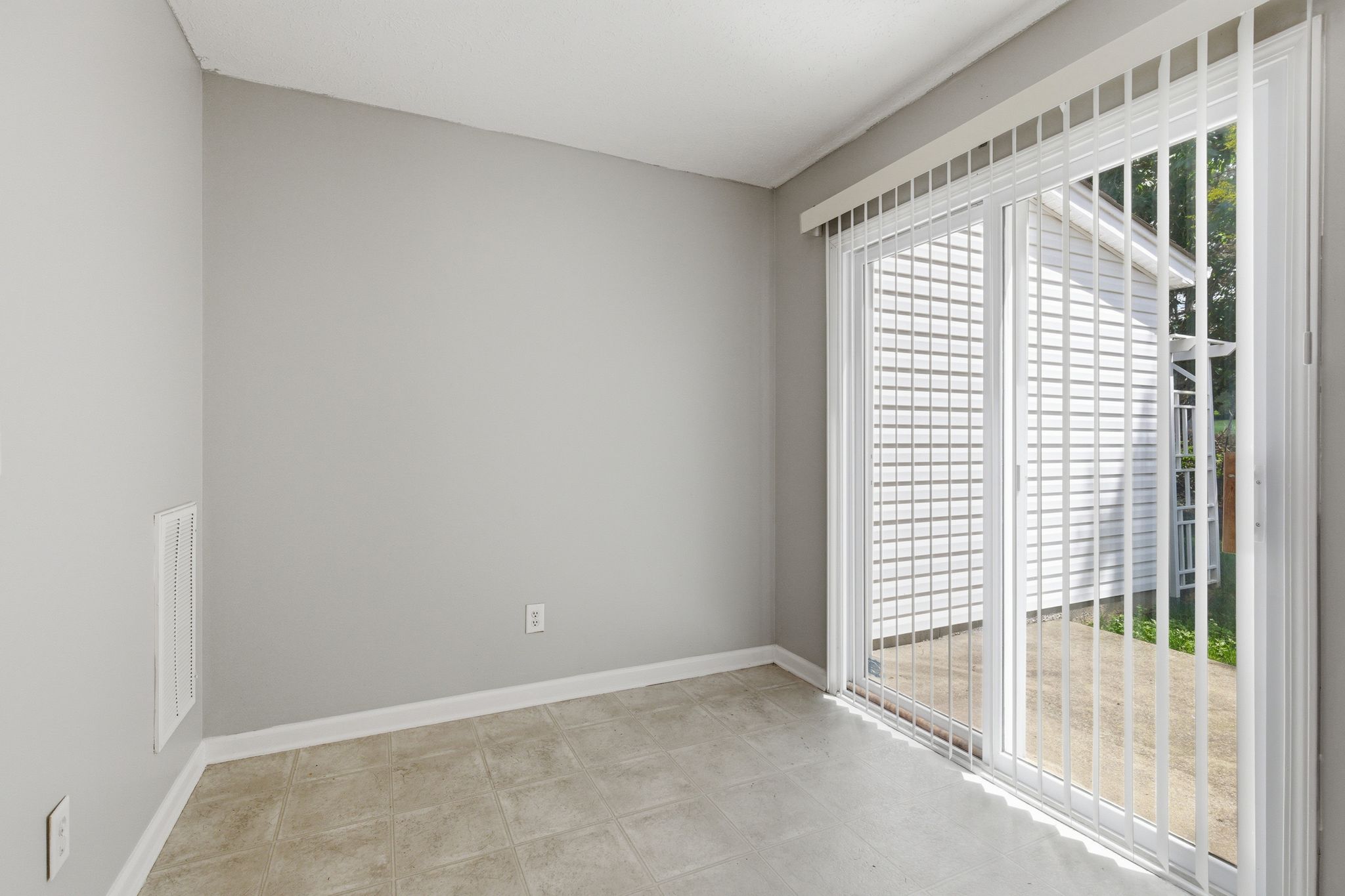 Empty sunroom with gray walls, light tiled floor, and large sliding glass doors with vertical blinds opening to a backyard area outside.