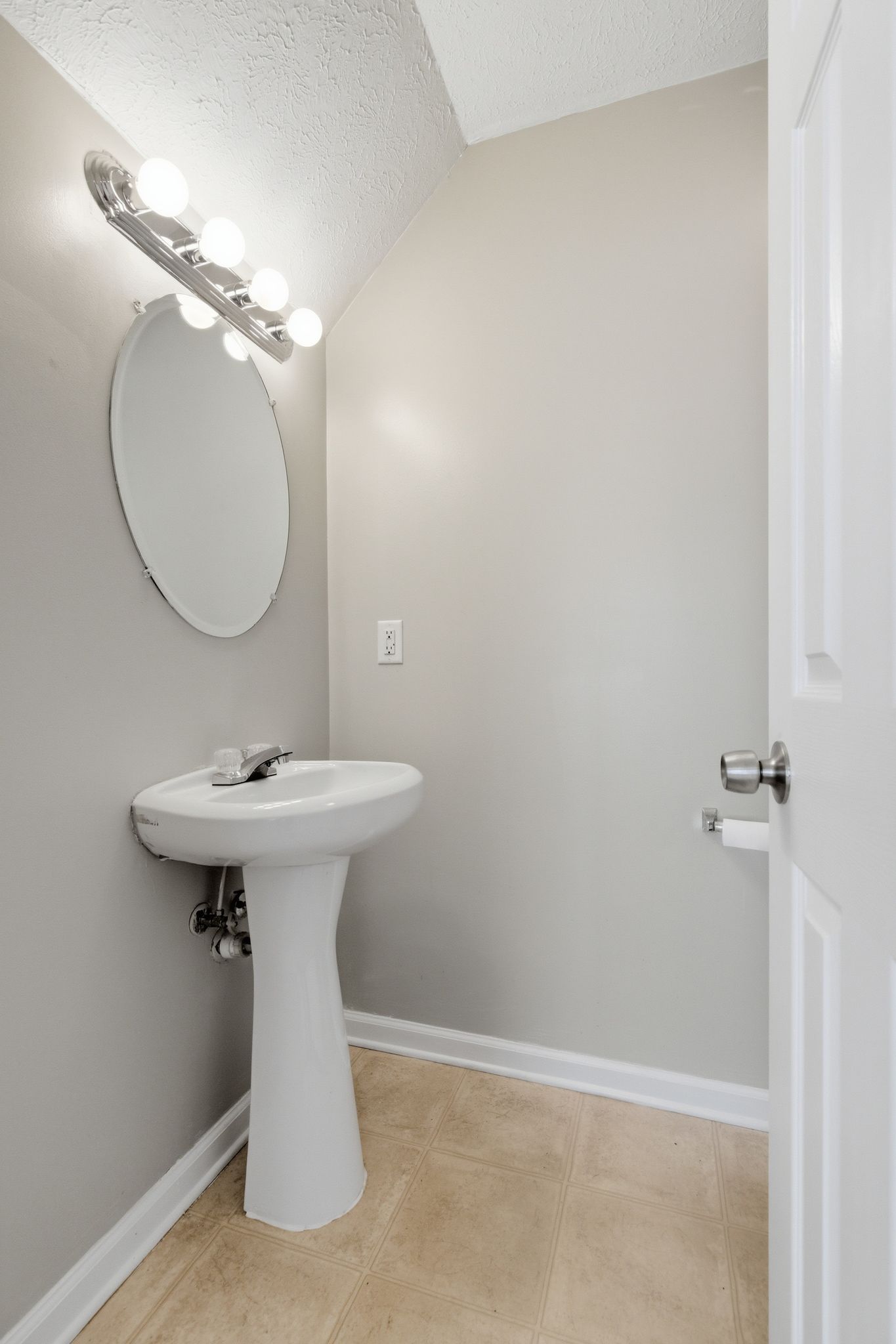 Small powder room with a pedestal sink, round mirror, and bright four-bulb vanity light above the sink on a gray wall.
