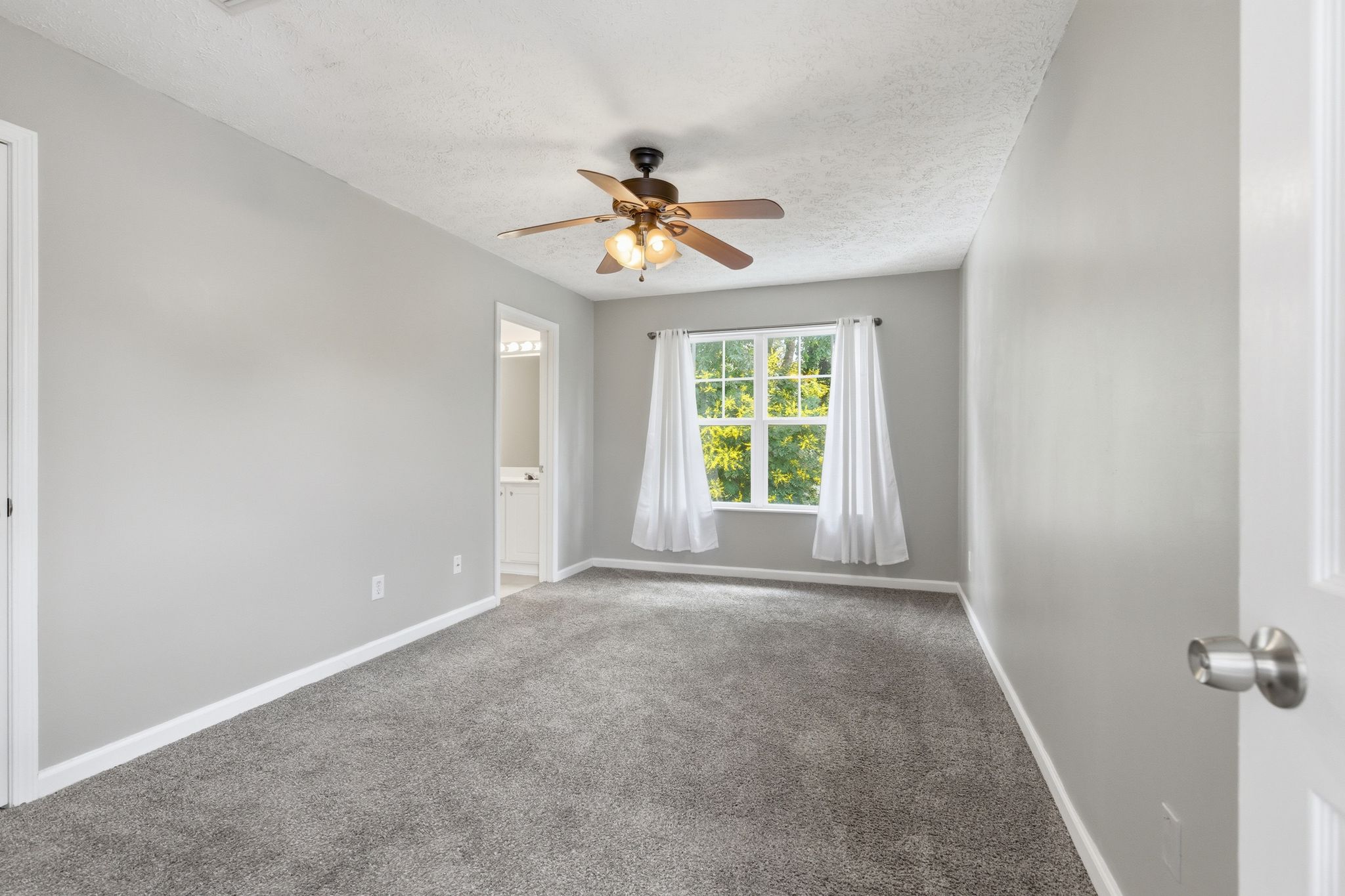 Empty gray bedroom with a ceiling fan, white curtains, and a window overlooking greenery outside the room