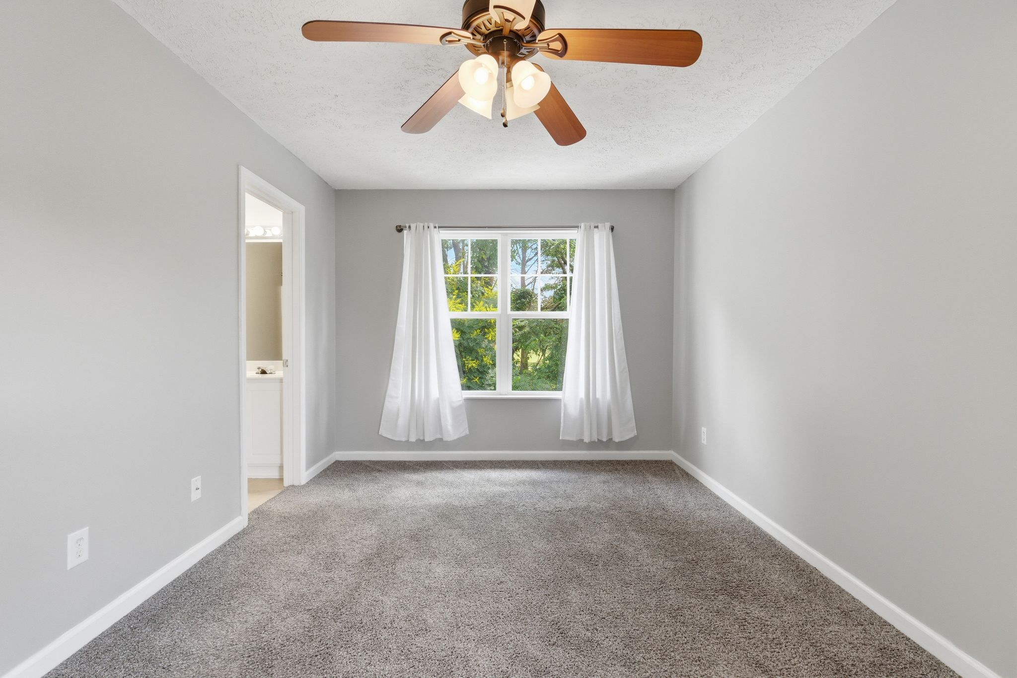 Light gray empty bedroom with a window and white curtains, carpeted floor, and a ceiling fan with wooden blades overhead