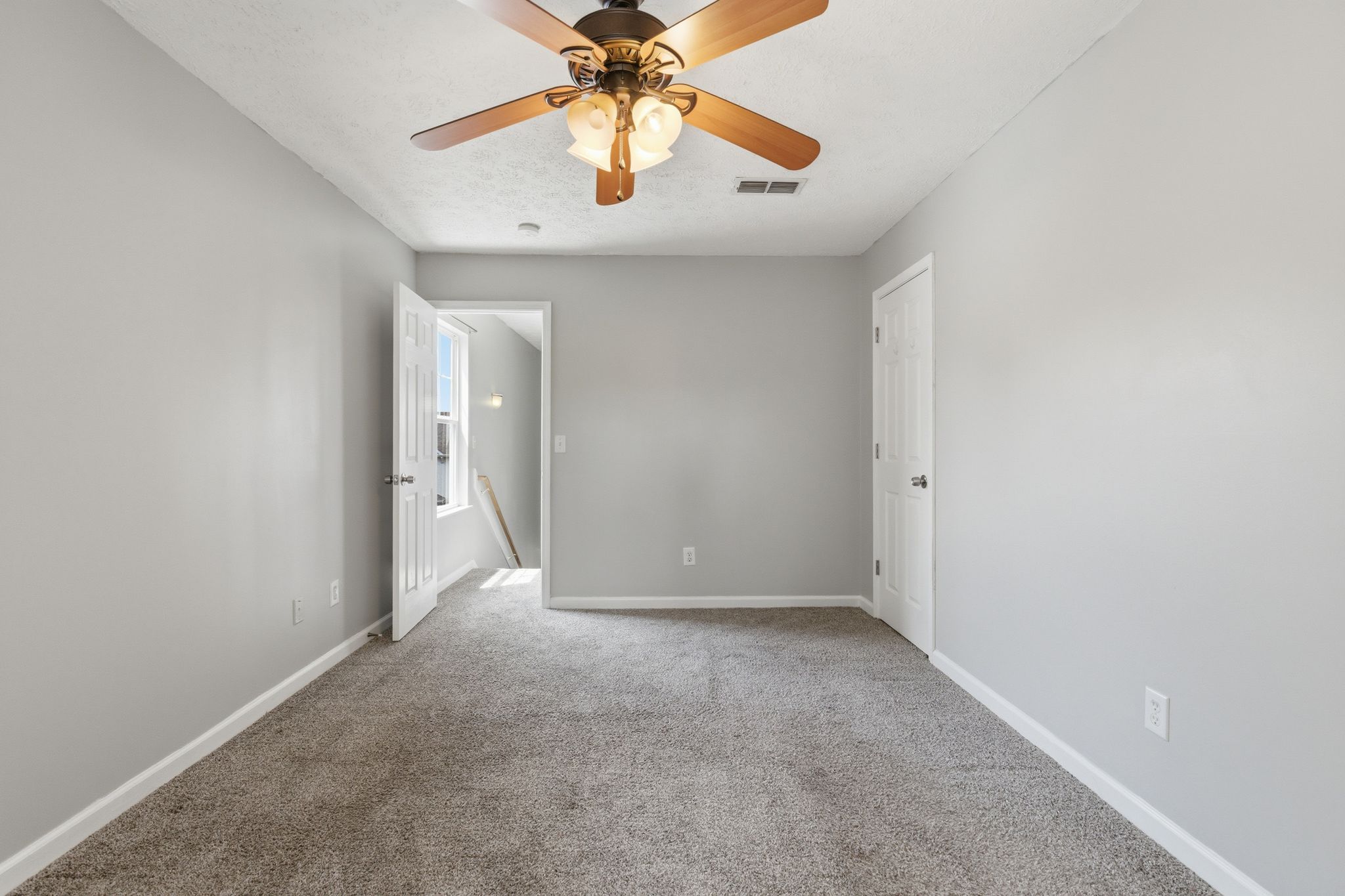 Empty gray carpeted room with two white doors, a ceiling fan, and natural light from a windowed doorway at the far end.