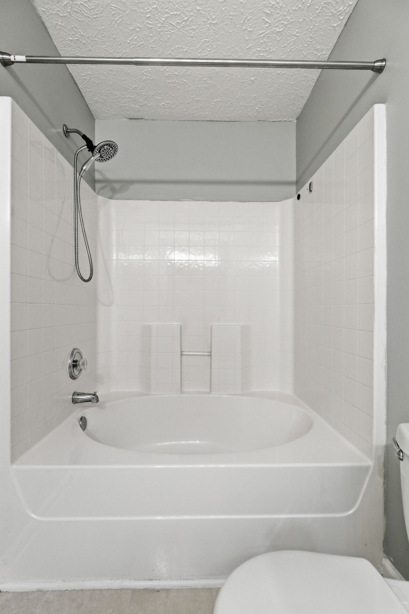White tiled bathtub with a handheld showerhead and flexible hose, a ceiling-mounted towel rod, and a partial view of a toilet in the foreground.