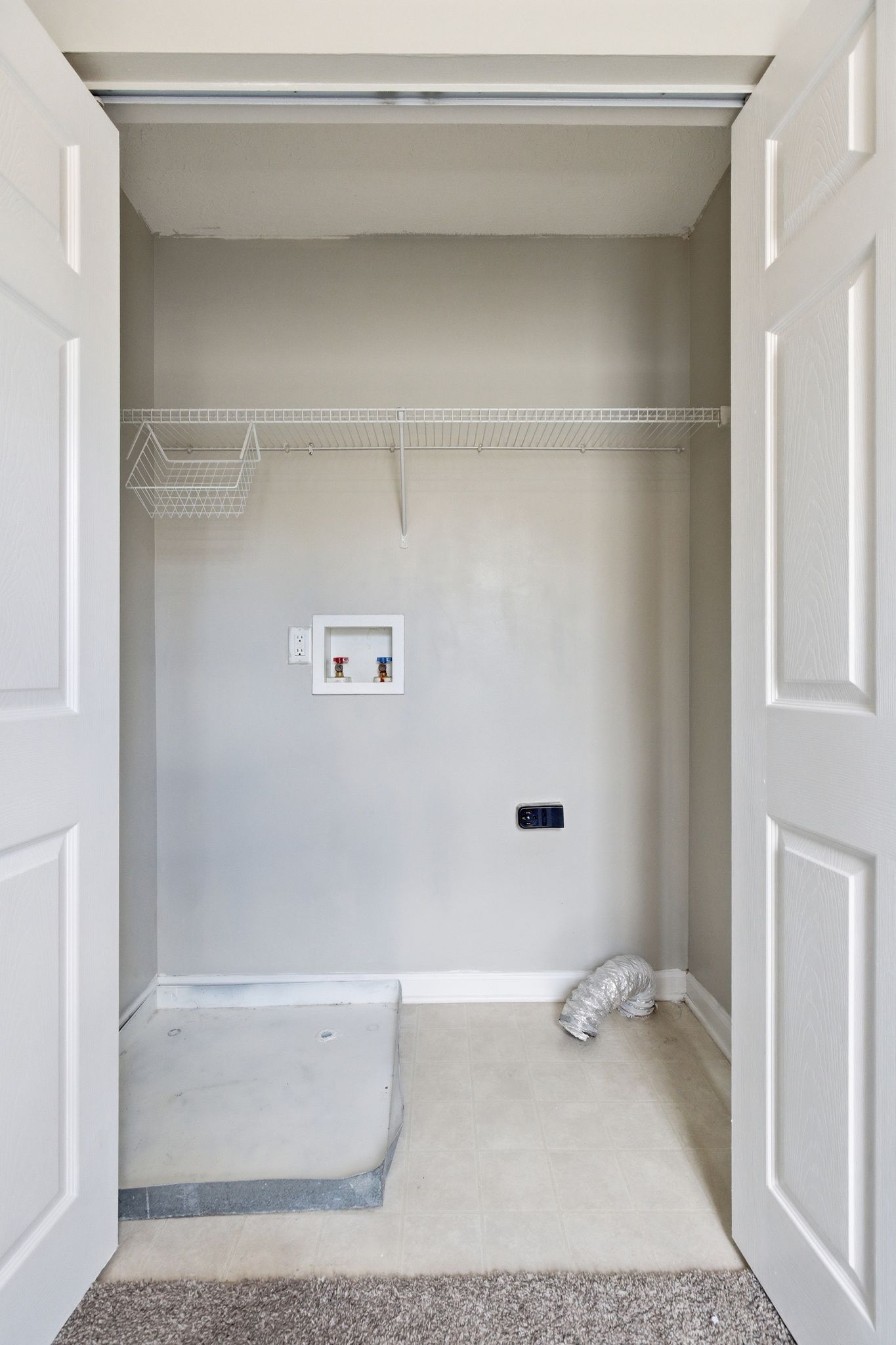 Small laundry closet with a white wire shelf, water-supply box, and loose vent hose on the tiled floor.