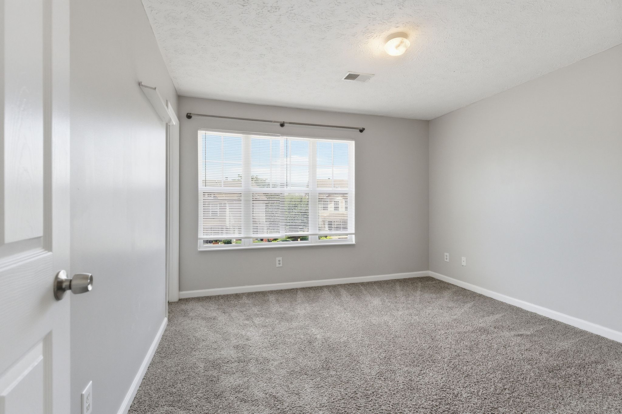 Empty light-gray room with a window covered by horizontal blinds, carpeted floor, and a ceiling light fixture; door partially visible on the left.