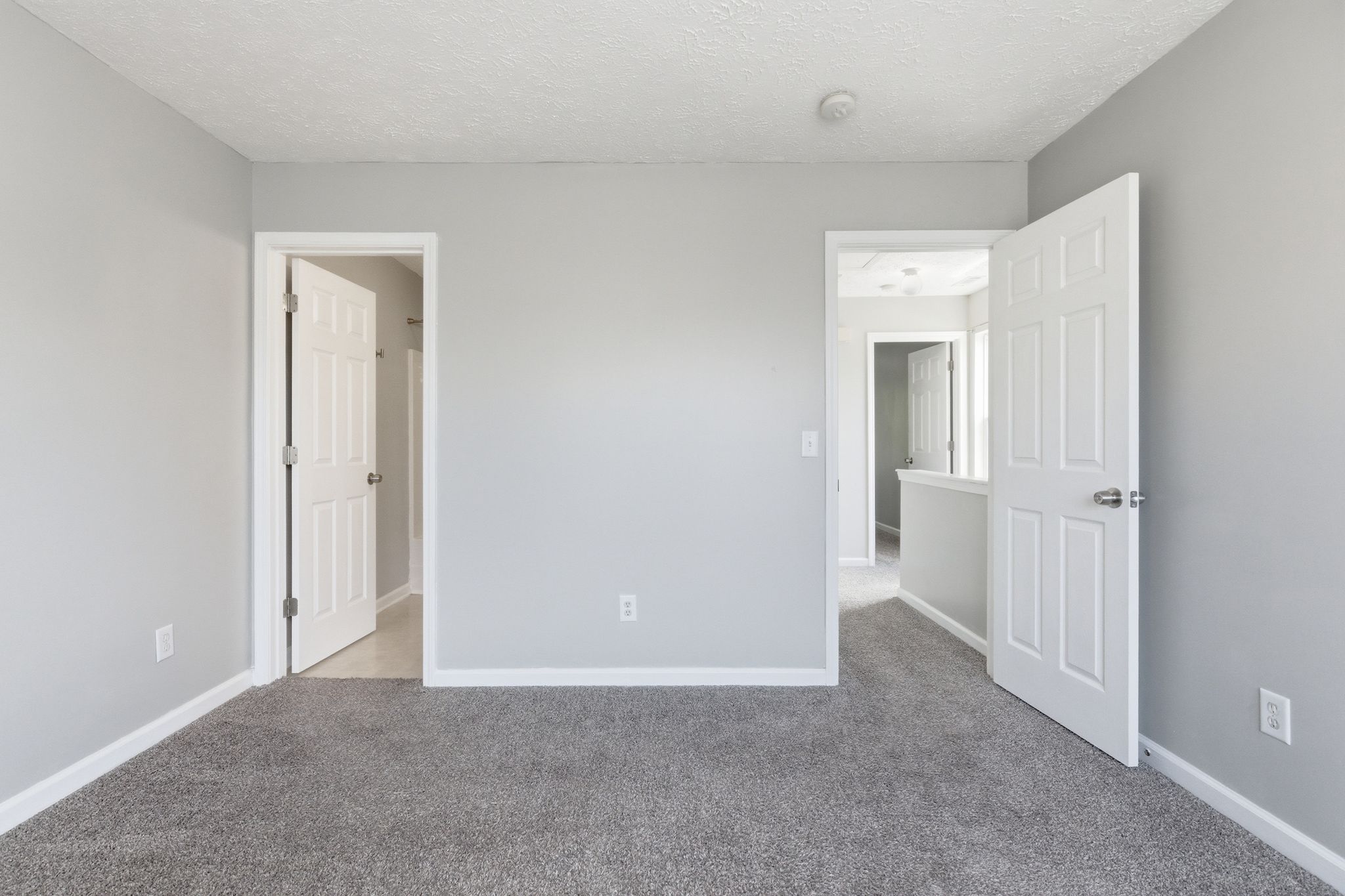 Empty light-gray bedroom with beige carpet and two white doors open to adjacent rooms, visible hallway and mirrors in the distance