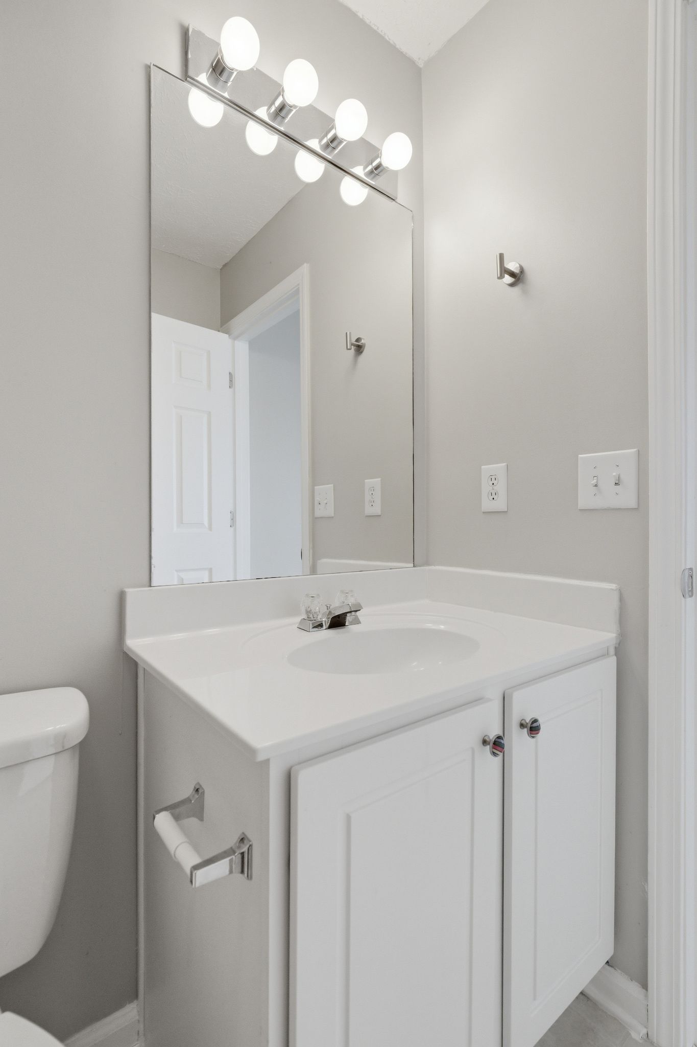 White bathroom vanity with a glossy sink, cabinet doors, and a large mirror framed by bright globe lights above the counter.