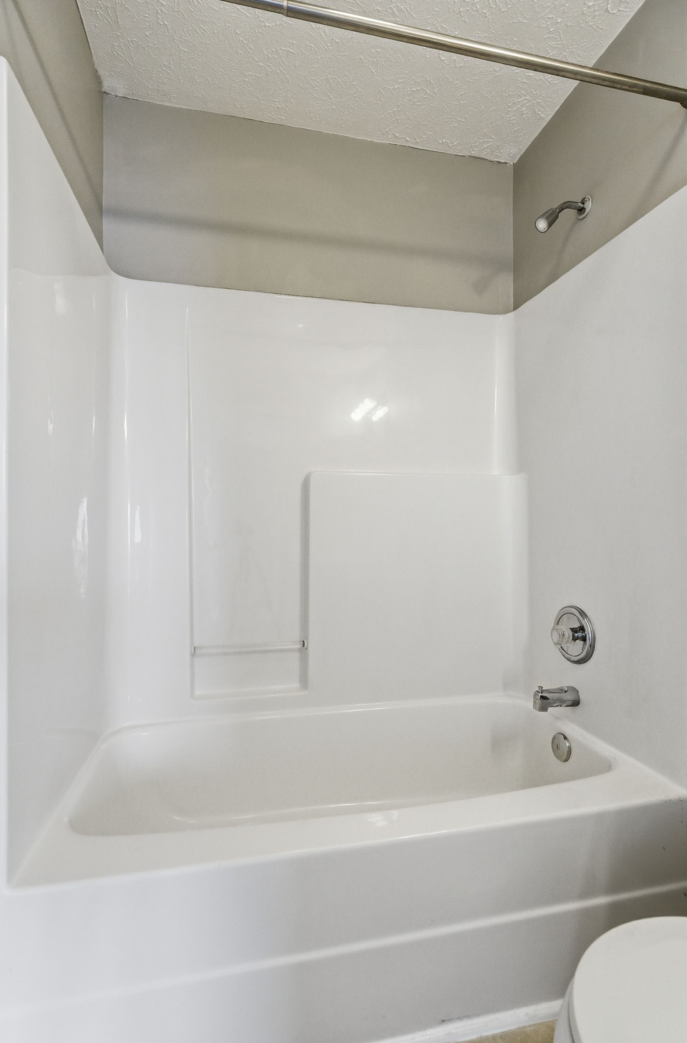 White bathtub with a built-in niche and chrome faucet/shower fixtures in a gray surround, metal curtain rod above, and textured ceiling.