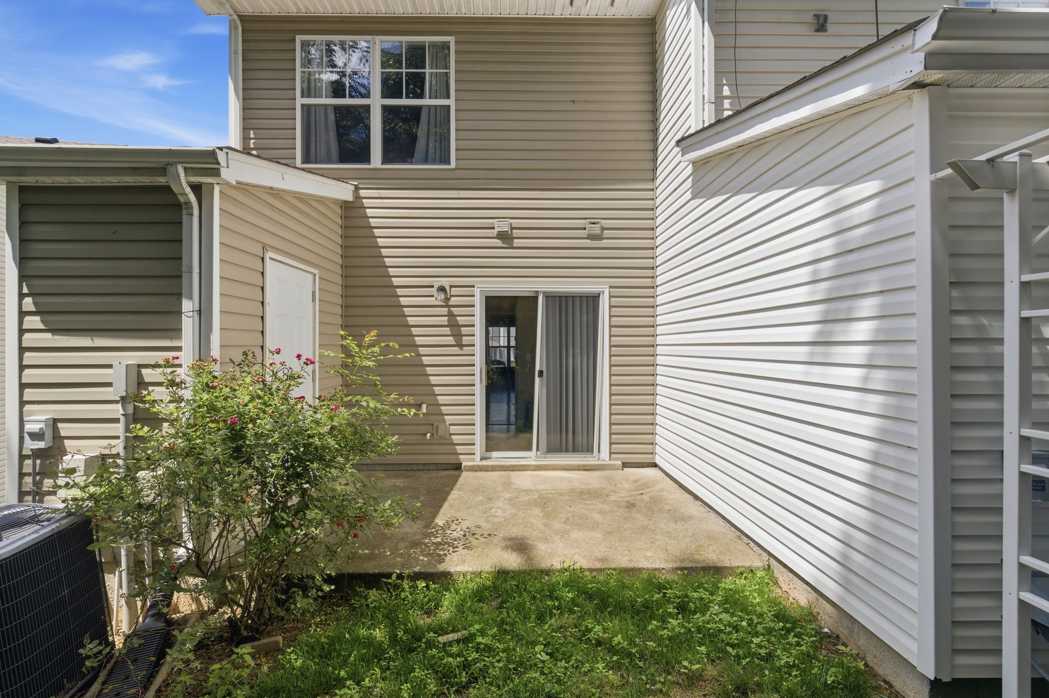 Backyard area between beige siding buildings with a sliding glass door, small patio, and overgrown grass and bushes under a clear blue sky.
