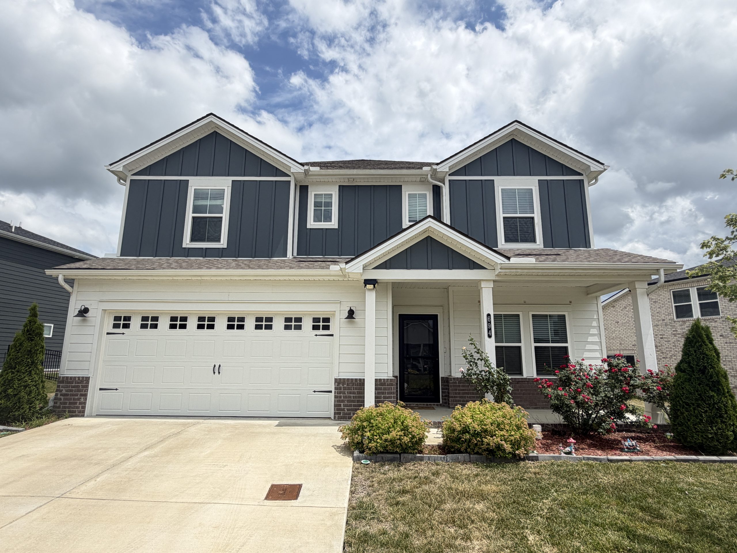 Two-story blue suburban house with a white garage door and a covered entry, blue siding, and a manicured front yard under a partly cloudy sky