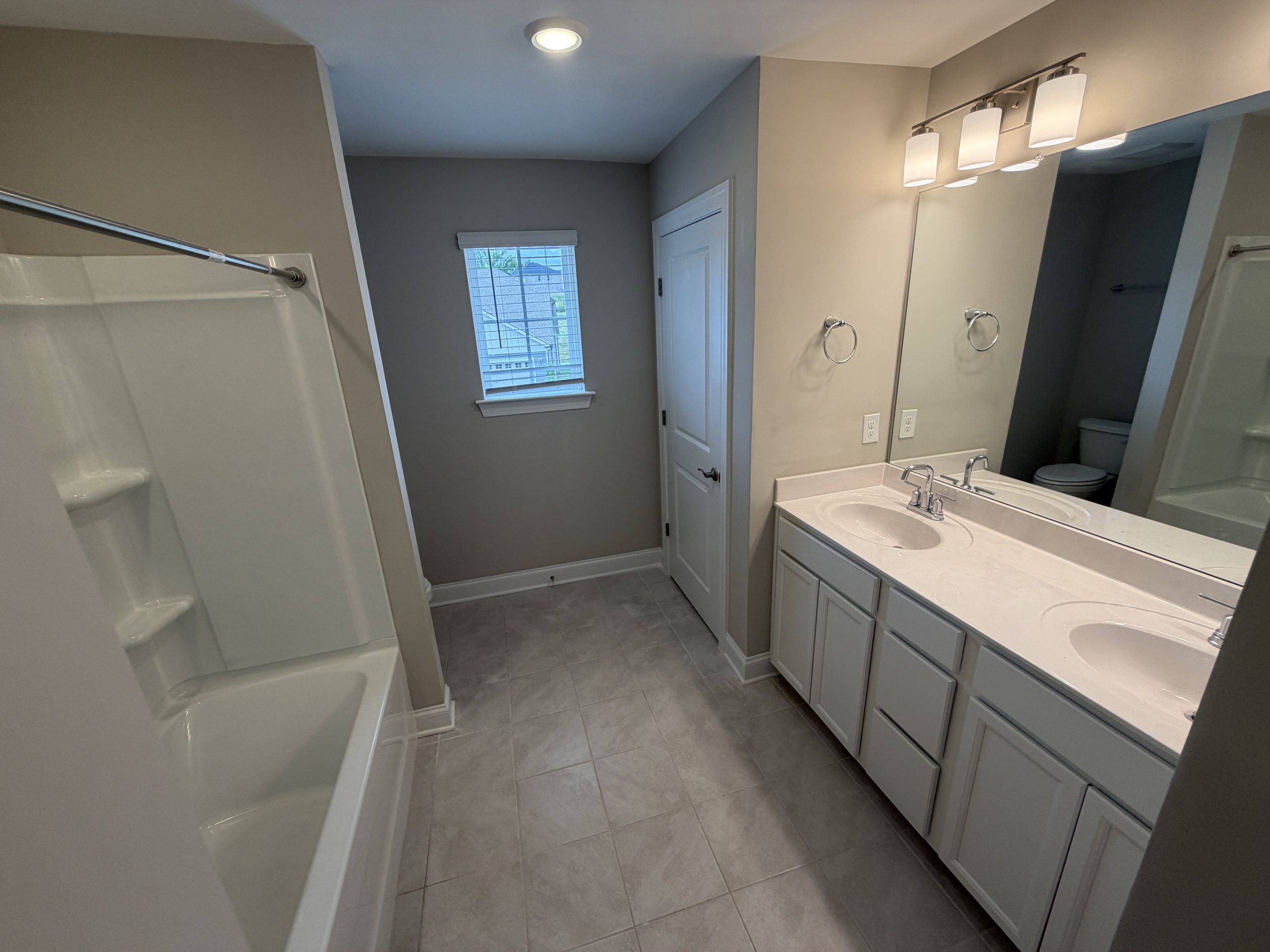 Double-sink bathroom vanity with a large mirror, bright light fixture, and a bathtub on the left; toilet visible in the back corner.