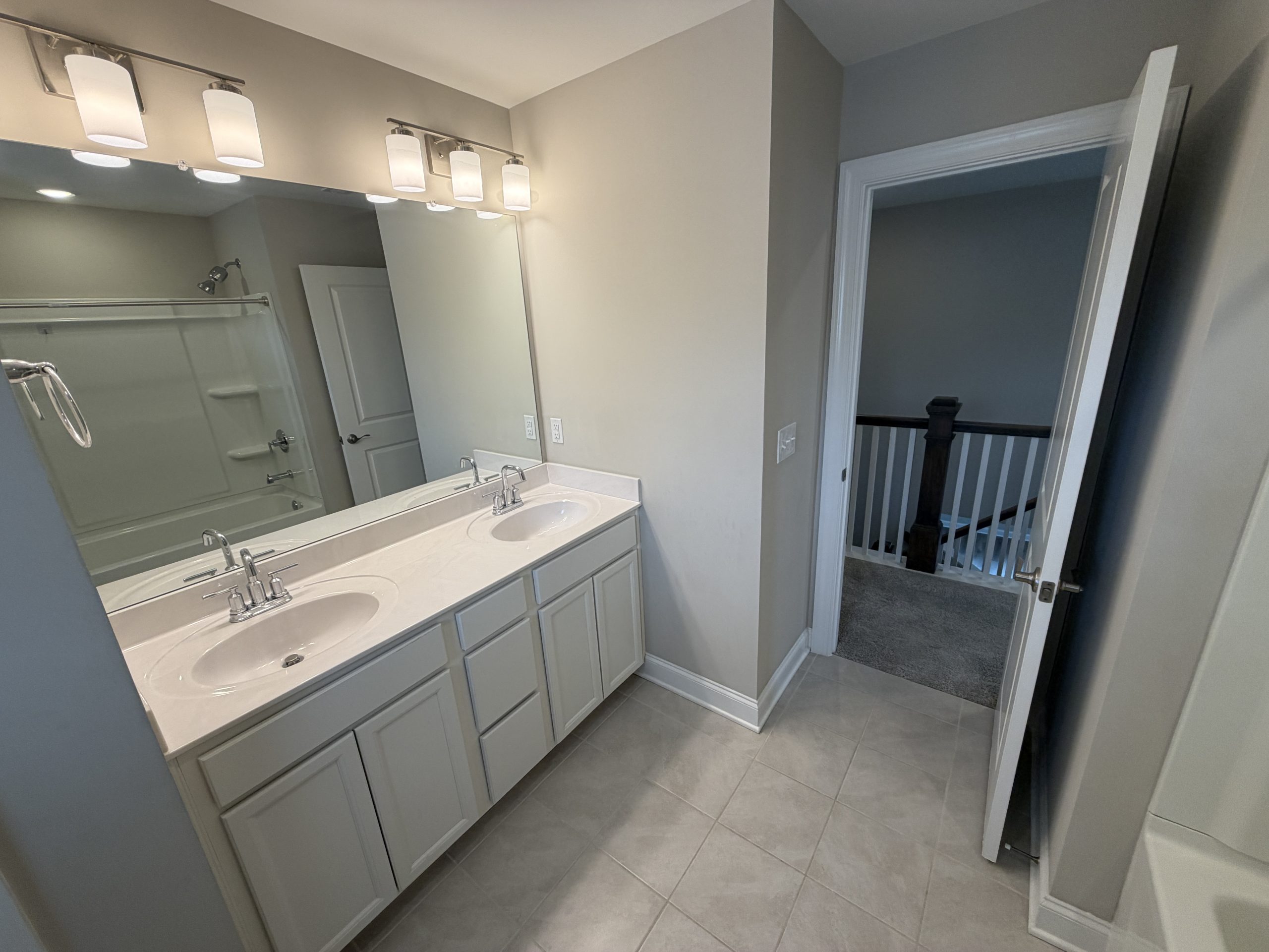 Modern bathroom with a double-sink vanity, large mirror, and quartet of ceiling lights above; shower/tub to the left and an open doorway to a hallway.