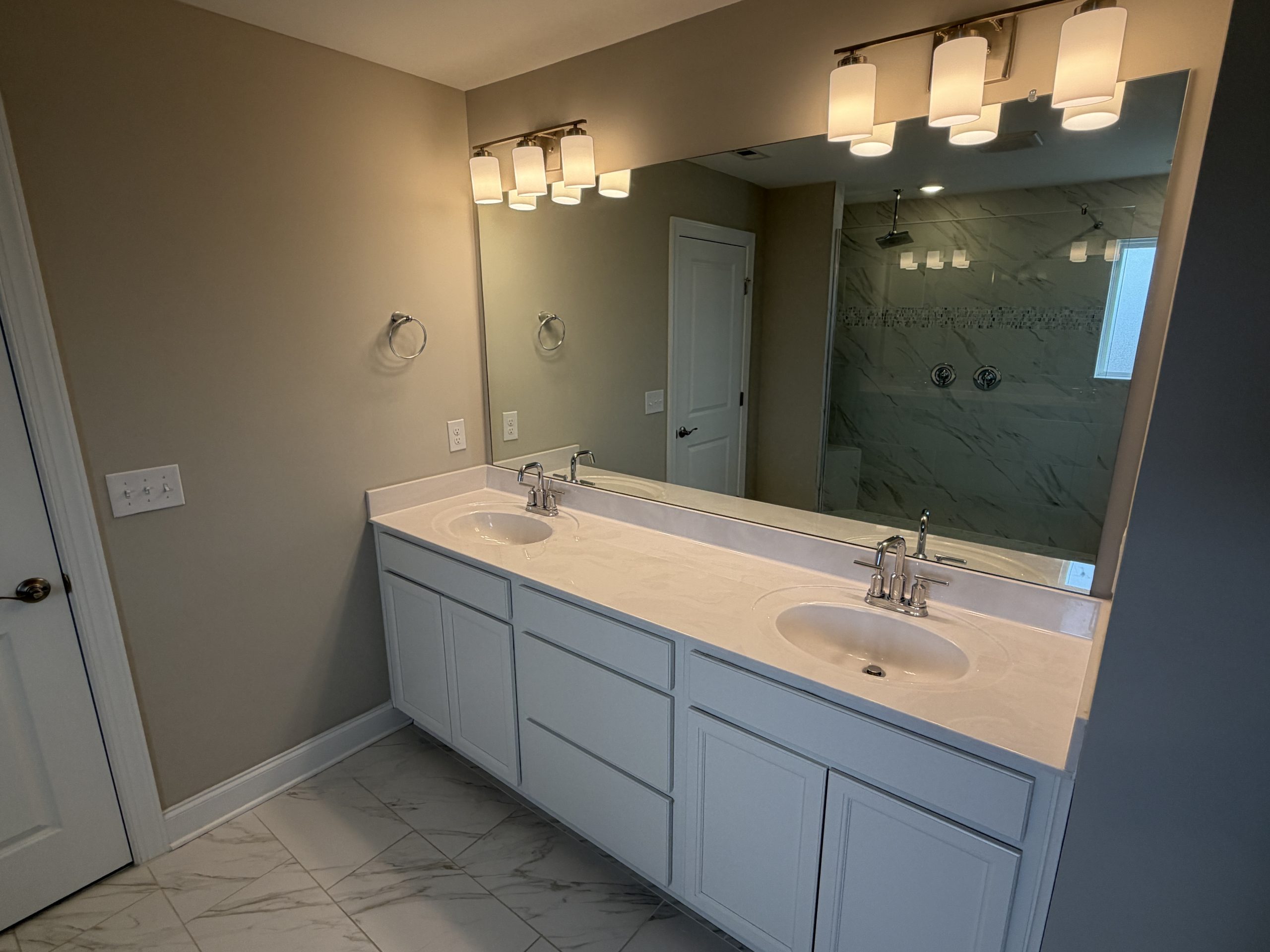 Double-sink bathroom vanity with white cabinets, large mirror, and modern wall lights above; glass-enclosed shower with marble-pattern walls in the background