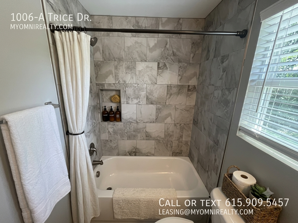 Marble-tiled bathroom with a white bathtub, shower curtain, and built-in niche holding bath products; window with blinds to the right and a towel rack on the left paneled wall.