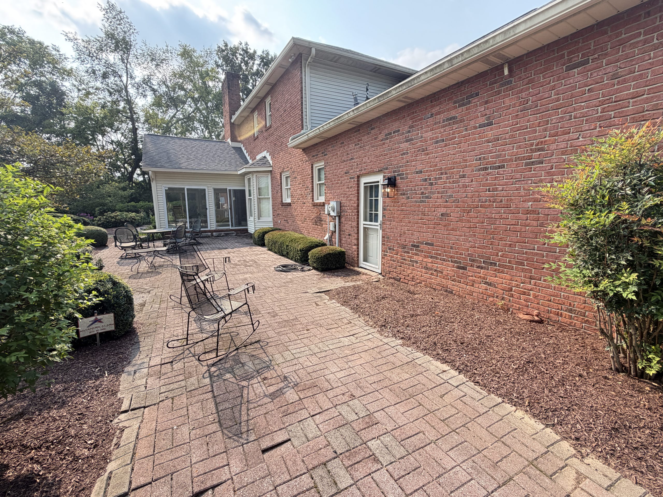 Brick house with an outdoor brick patio and black metal tables and chairs; screened porch on the left, trees in the background.