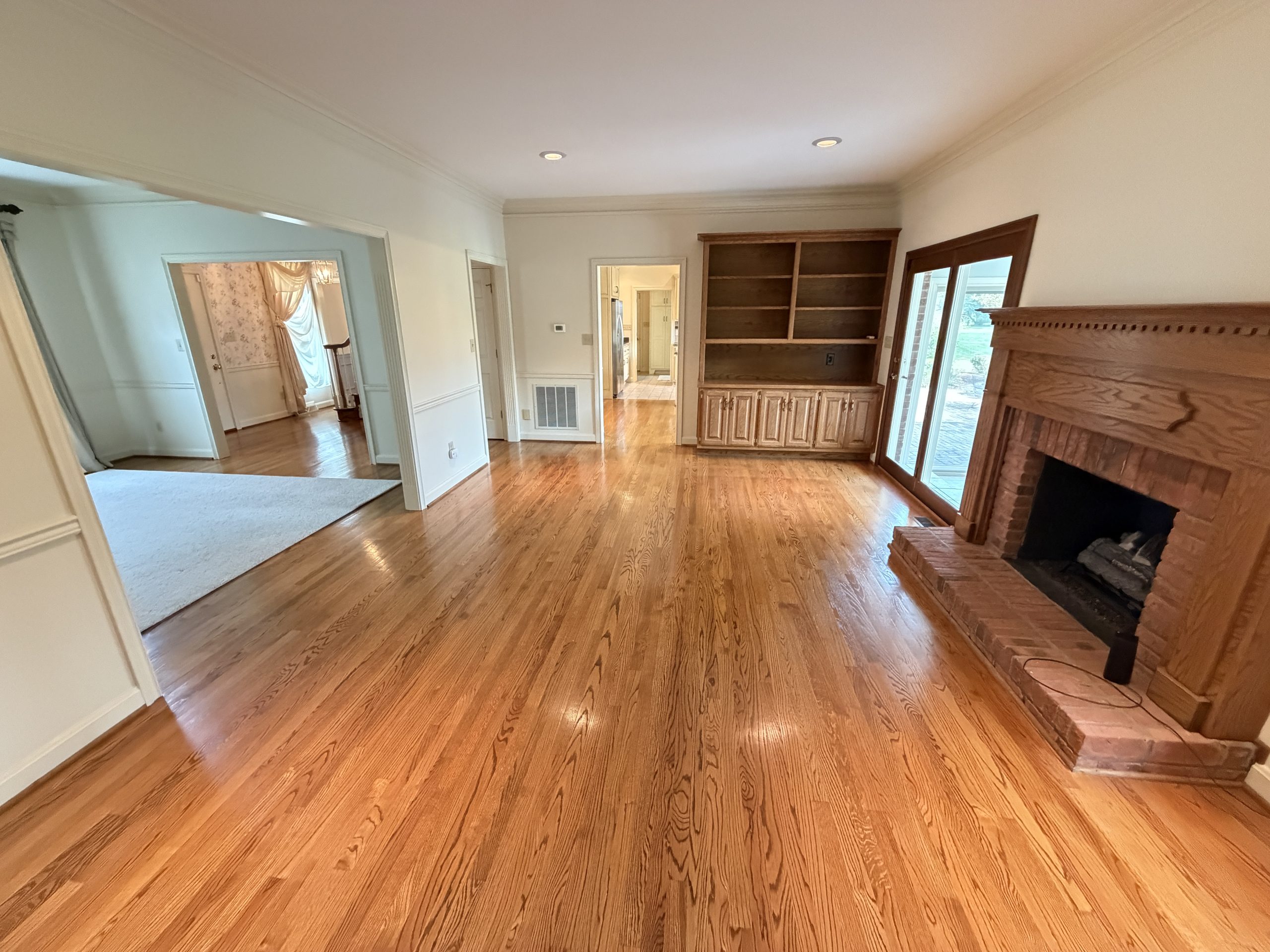 Spacious living room with oak hardwood floors, built-in shelves, and a brick fireplace on the right. French doors lead outside nearby.