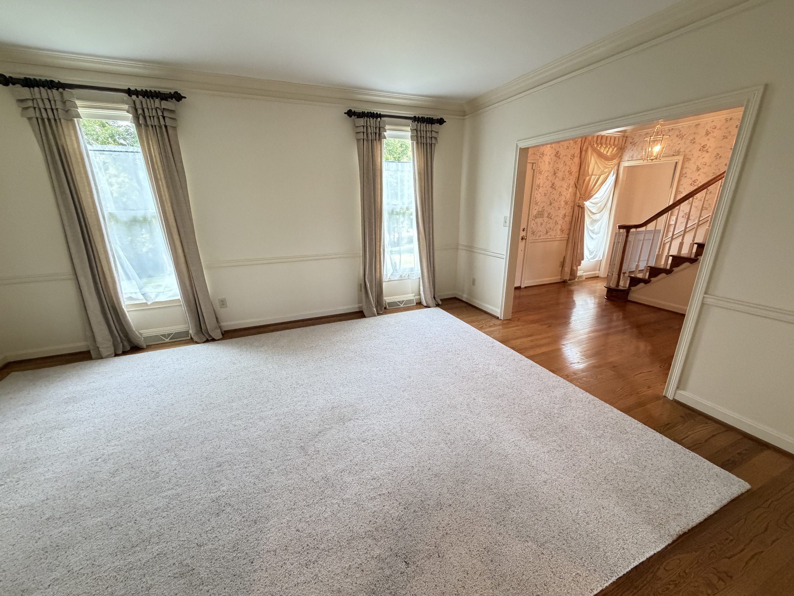 Living room with light carpet, two tall windows with gray curtains, and white walls. A doorway leads to a staircase area with floral wallpaper.