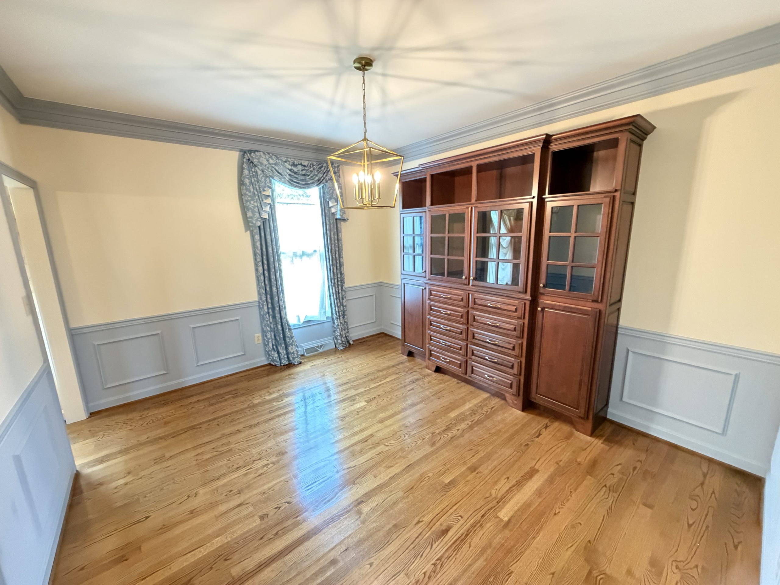 A dining room with a tall wooden china cabinet, chandelier, and blue patterned curtains by a window, hardwood floor, and wainscoted walls.