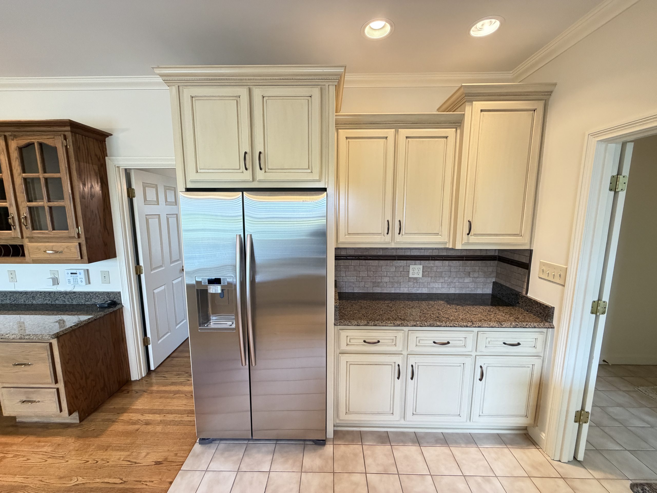 Kitchen with light beige raised-panel cabinets surrounding a stainless steel side-by-side refrigerator; tile floor and granite counters visible on left and right edges.