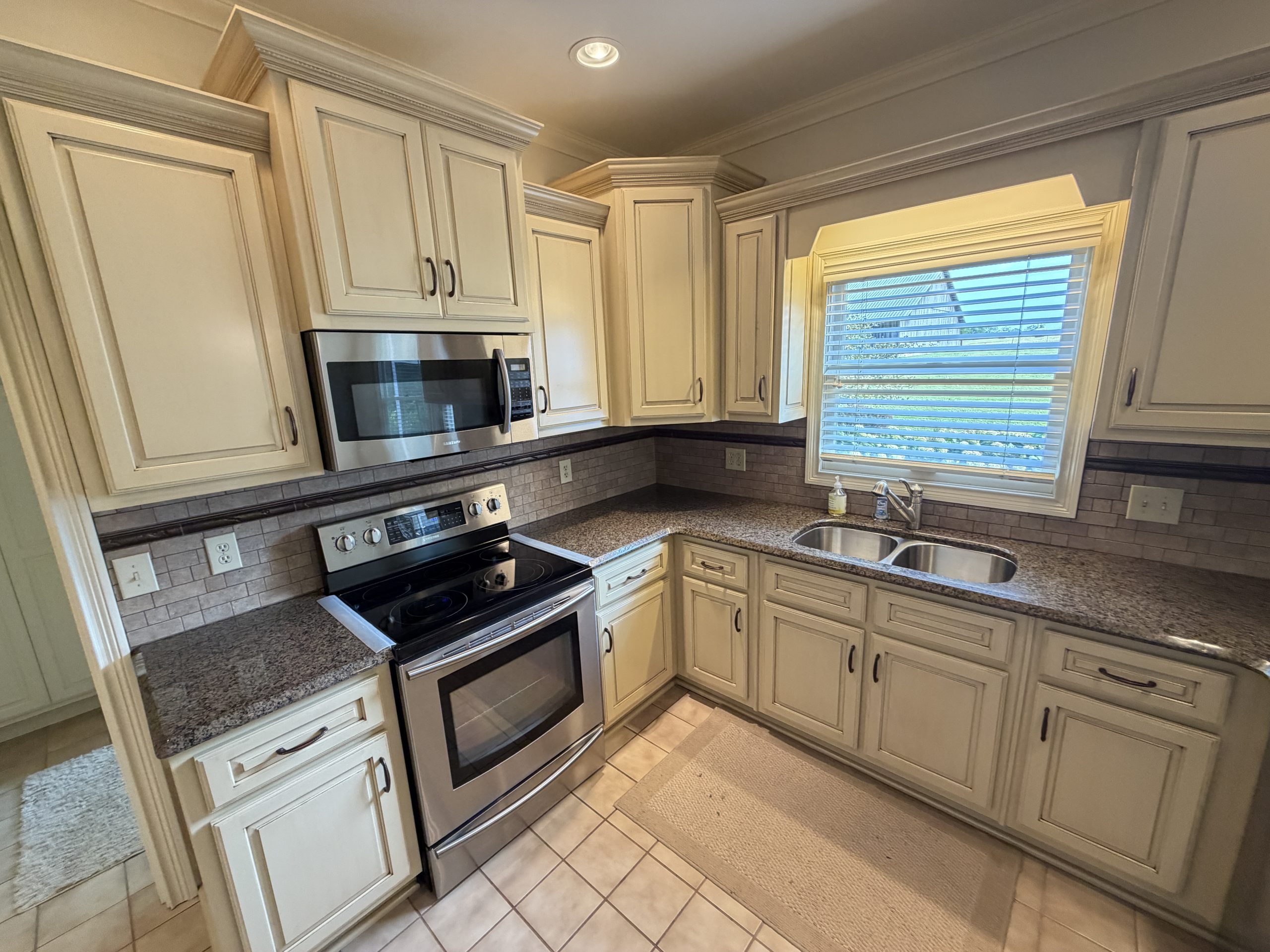 L-shaped kitchen with white raised-panel cabinets, granite countertops, and stainless steel stove and microwave under a window with blinds.