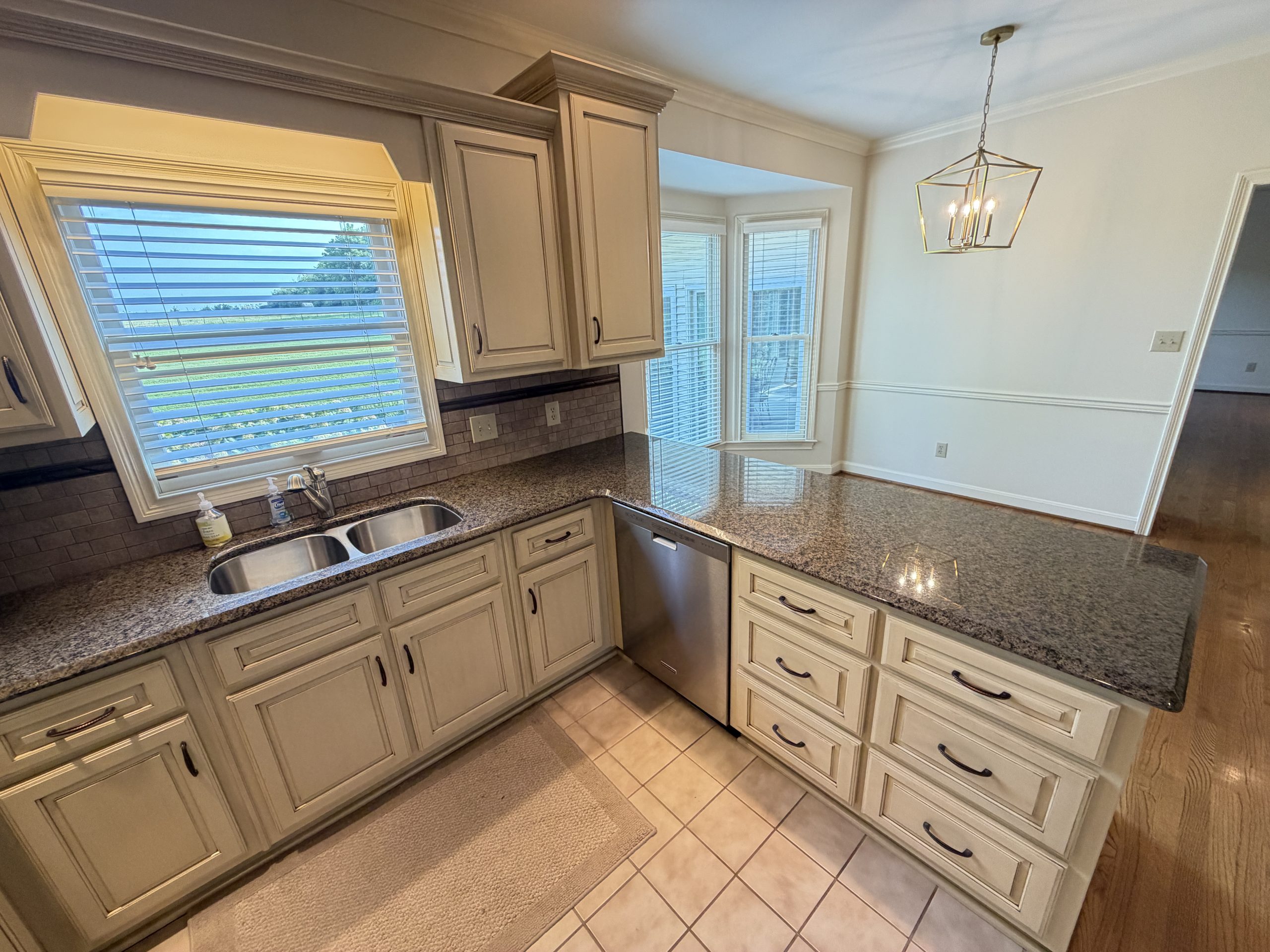 View of a bright U-shaped kitchen with white cabinets, granite counters, and a double sink beneath large windows with blinds