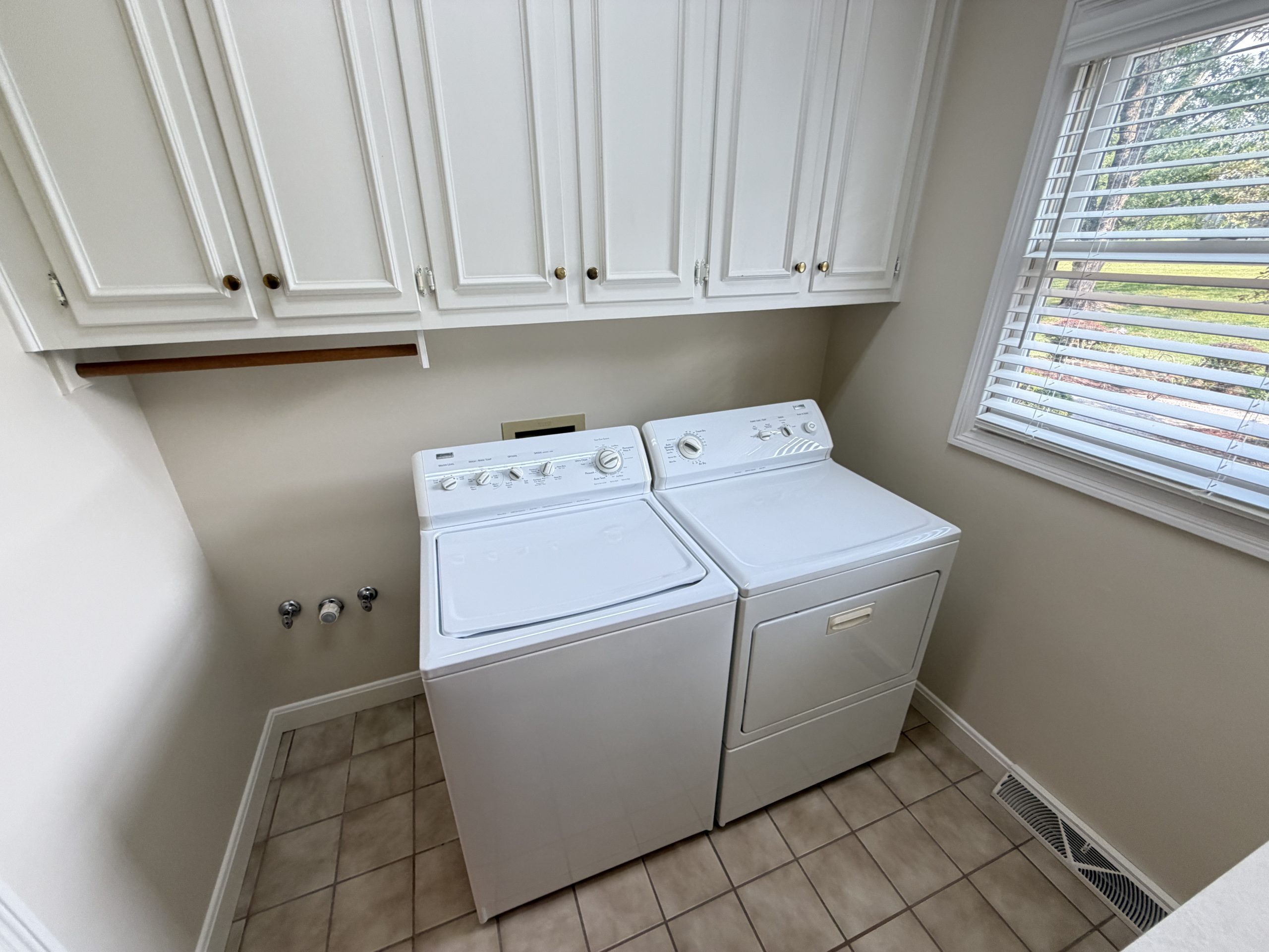 Laundry room with white cabinets above a stacked washer and dryer, a wooden shelf beneath the cabinets, and a window with blinds to the right.