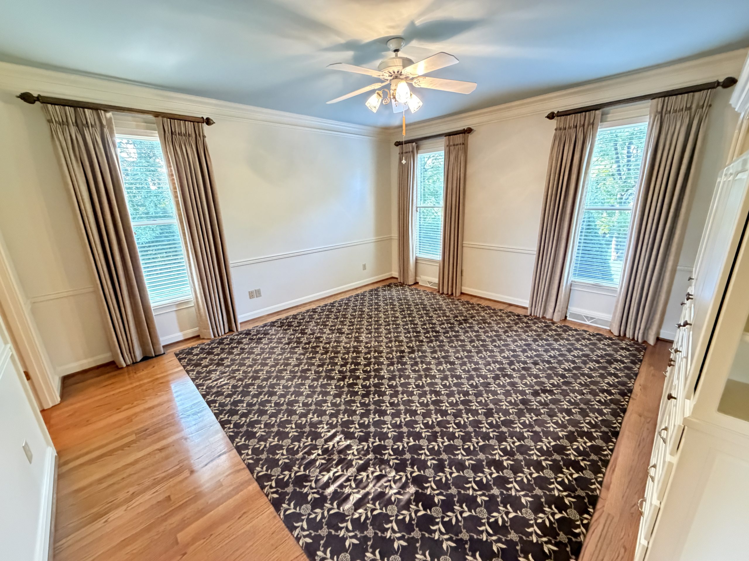 Sunlit living room with four tall windows, beige curtains, white trim, a ceiling fan, and a patterned dark rug on wood floors.