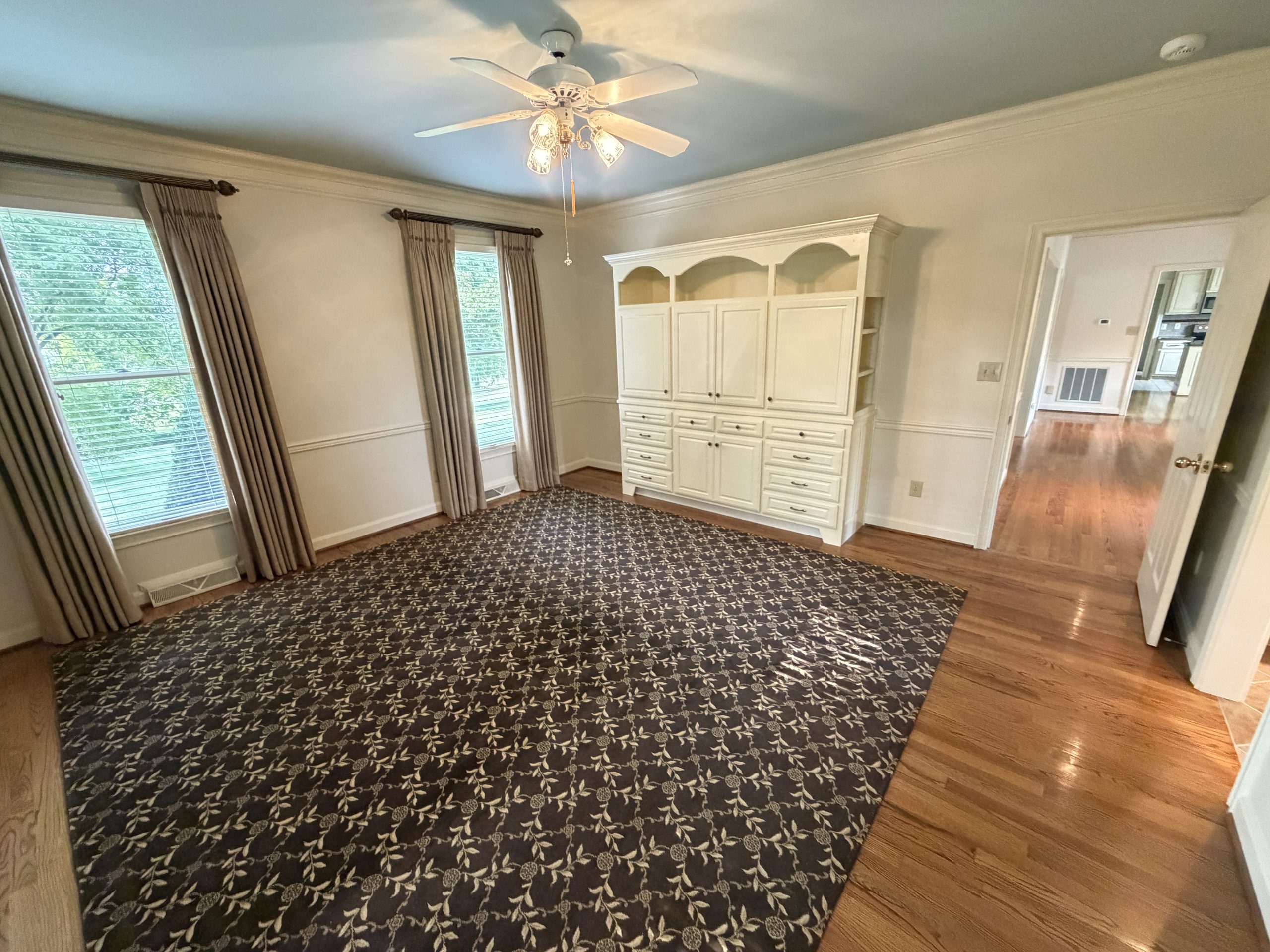 Bright foyer or living area with white built-in cabinet, dark patterned area rug, hardwood floors, and large windows with beige curtains; ceiling fan overhead; doorway to adjacent kitchen visible.