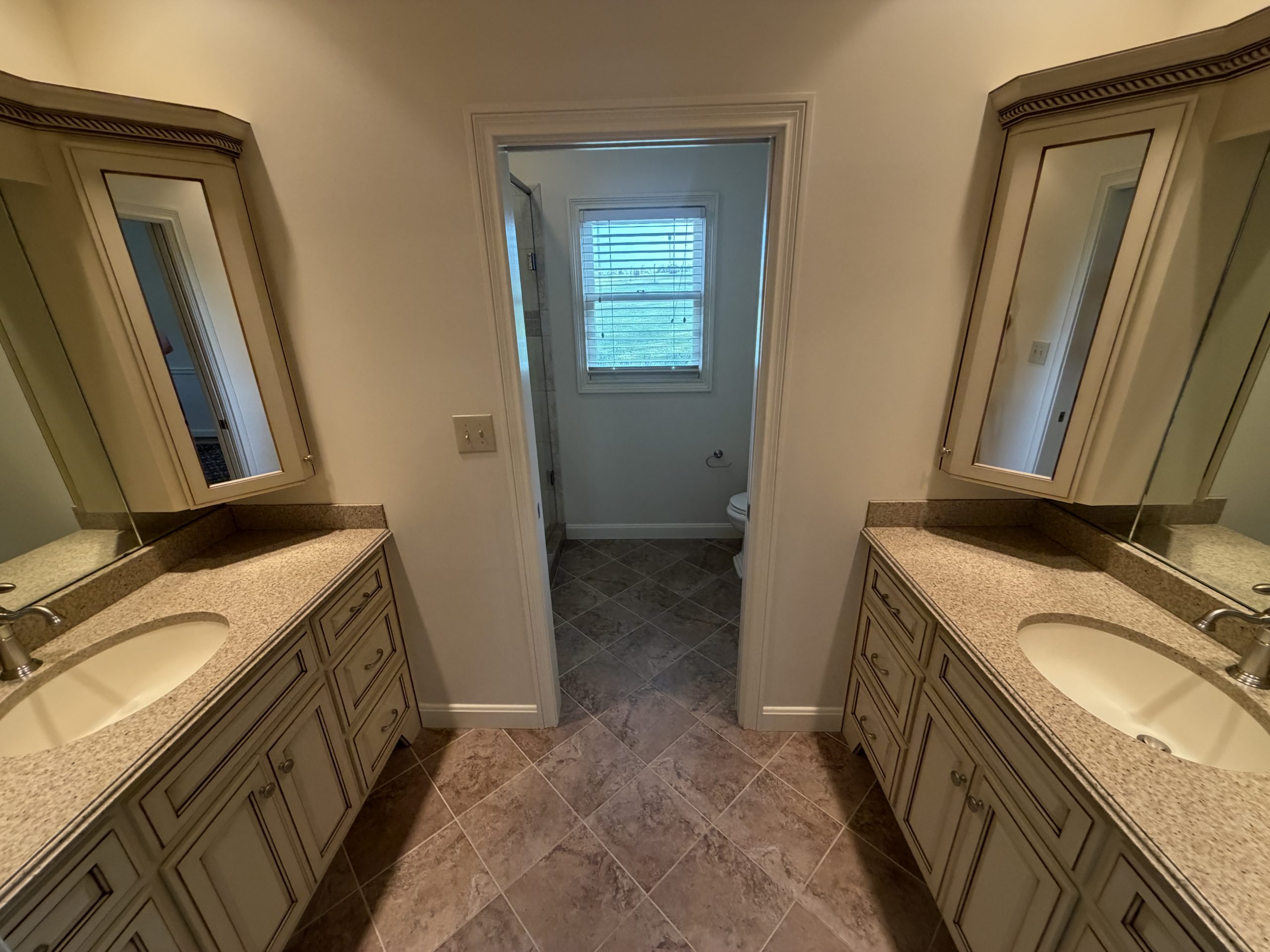 Bathroom with two mirrored vanities facing each other, granite countertops, and cream cabinets; doorway to a small room with a window and toilet ahead