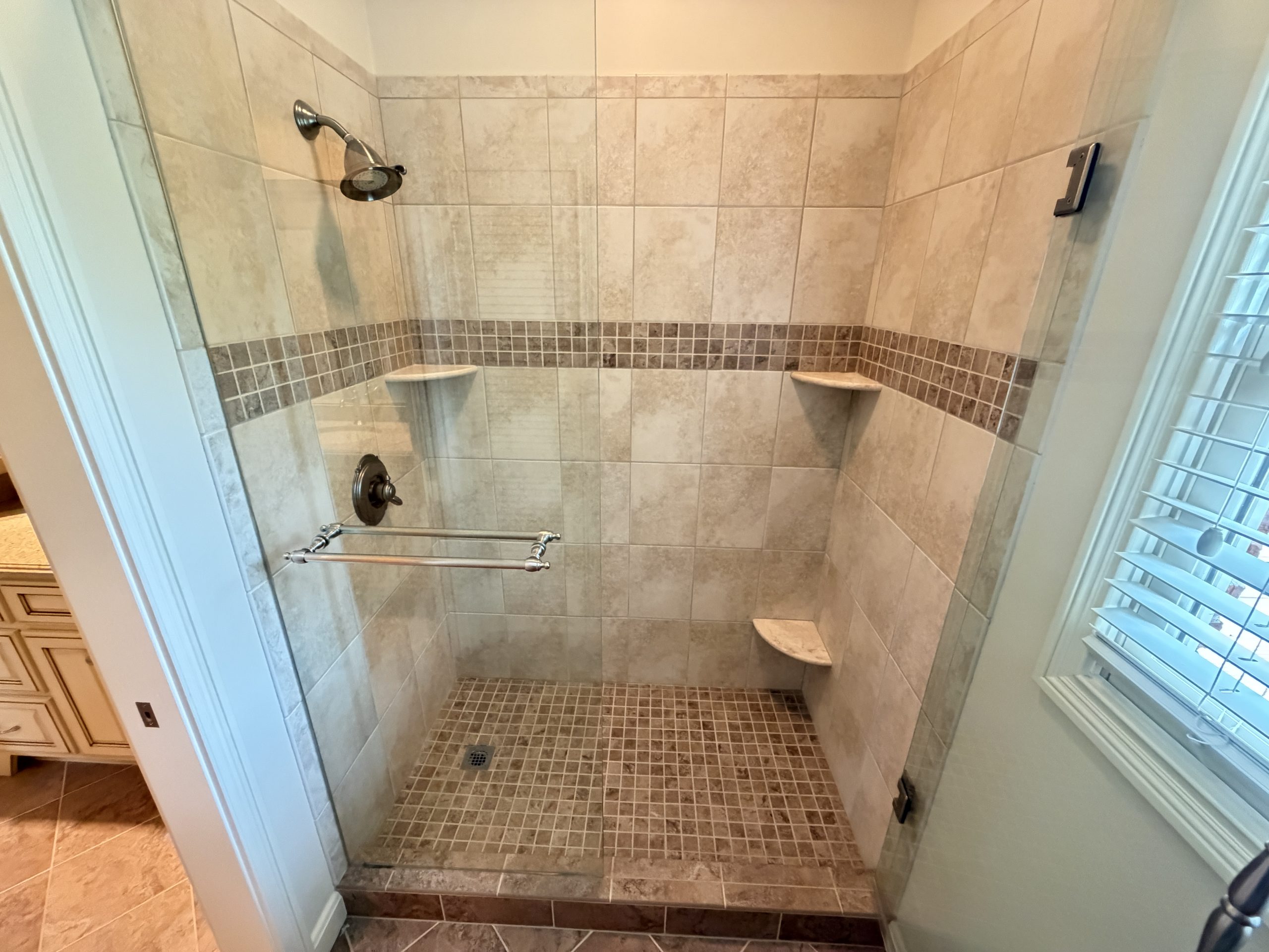 Walk-in tiled shower with glass door, a chrome showerhead, and two built-in corner shelves.