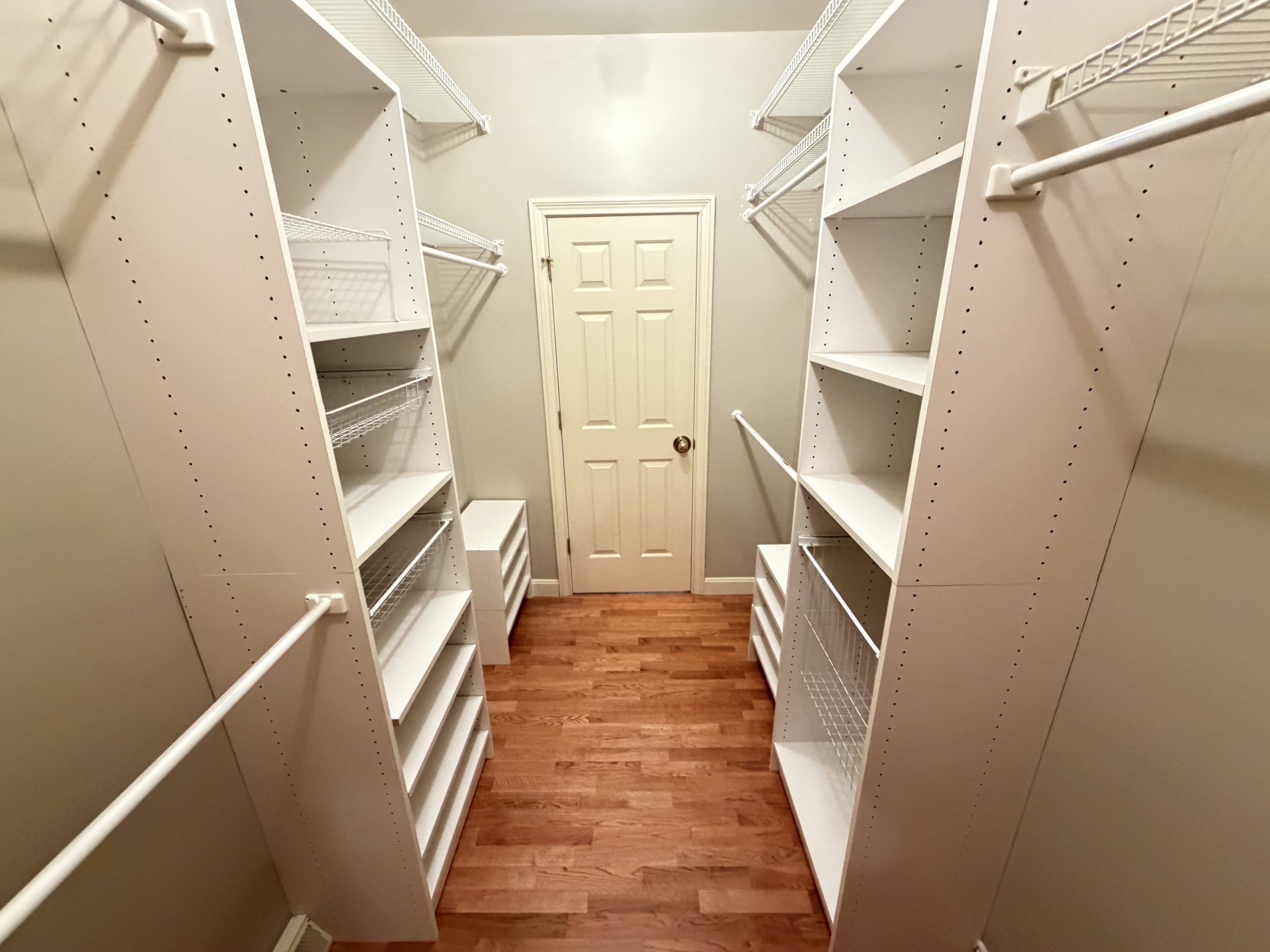 Narrow walk-in closet with white shelves, hanging rods, and a door at the far end.
