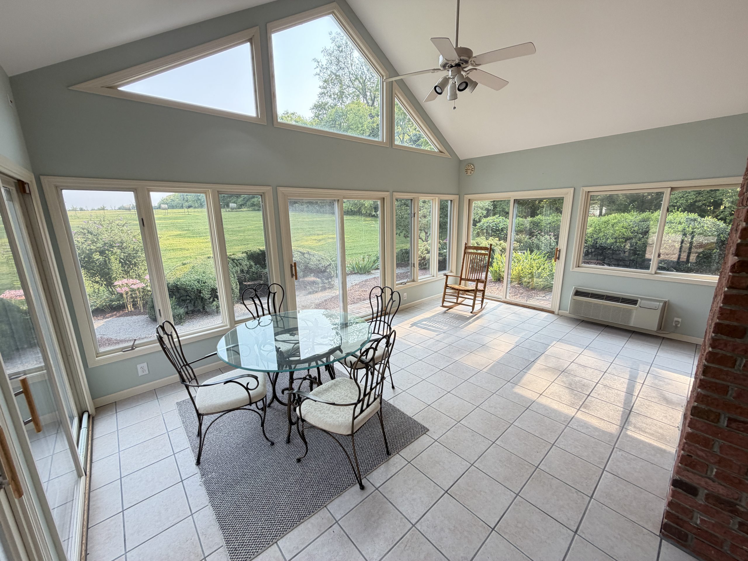 Bright sunroom with a round glass dining table, black wrought-iron chairs, and large windows overlooking a green garden behind.