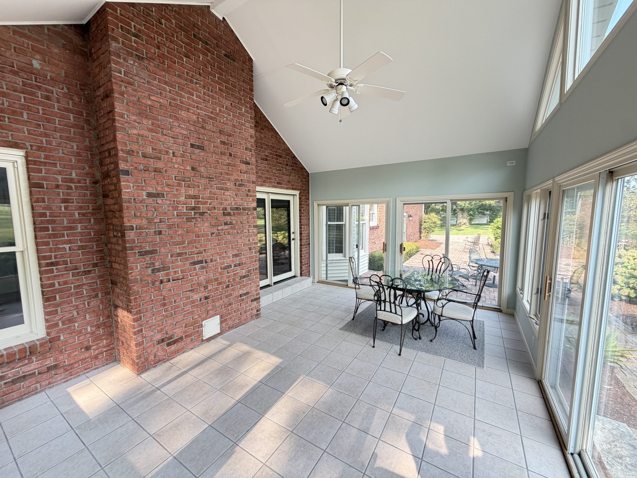 Enclosed sunroom with a brick wall, white ceiling fan, and glass dining table set on a tiled floor with large windows outside.