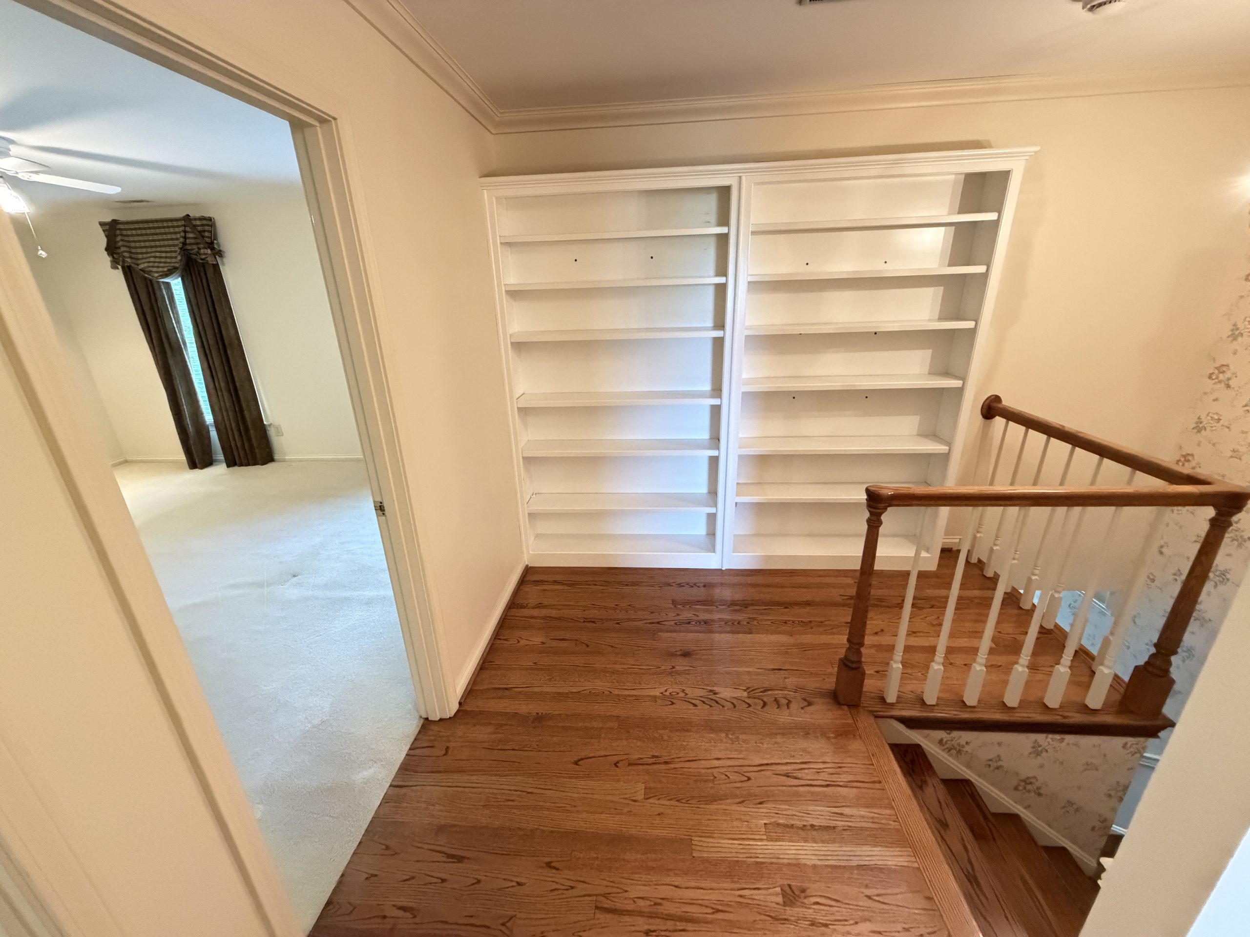 Upstairs hallway with a tall white built-in bookcase along the wall, wooden floor, and a wooden handrail on the right.