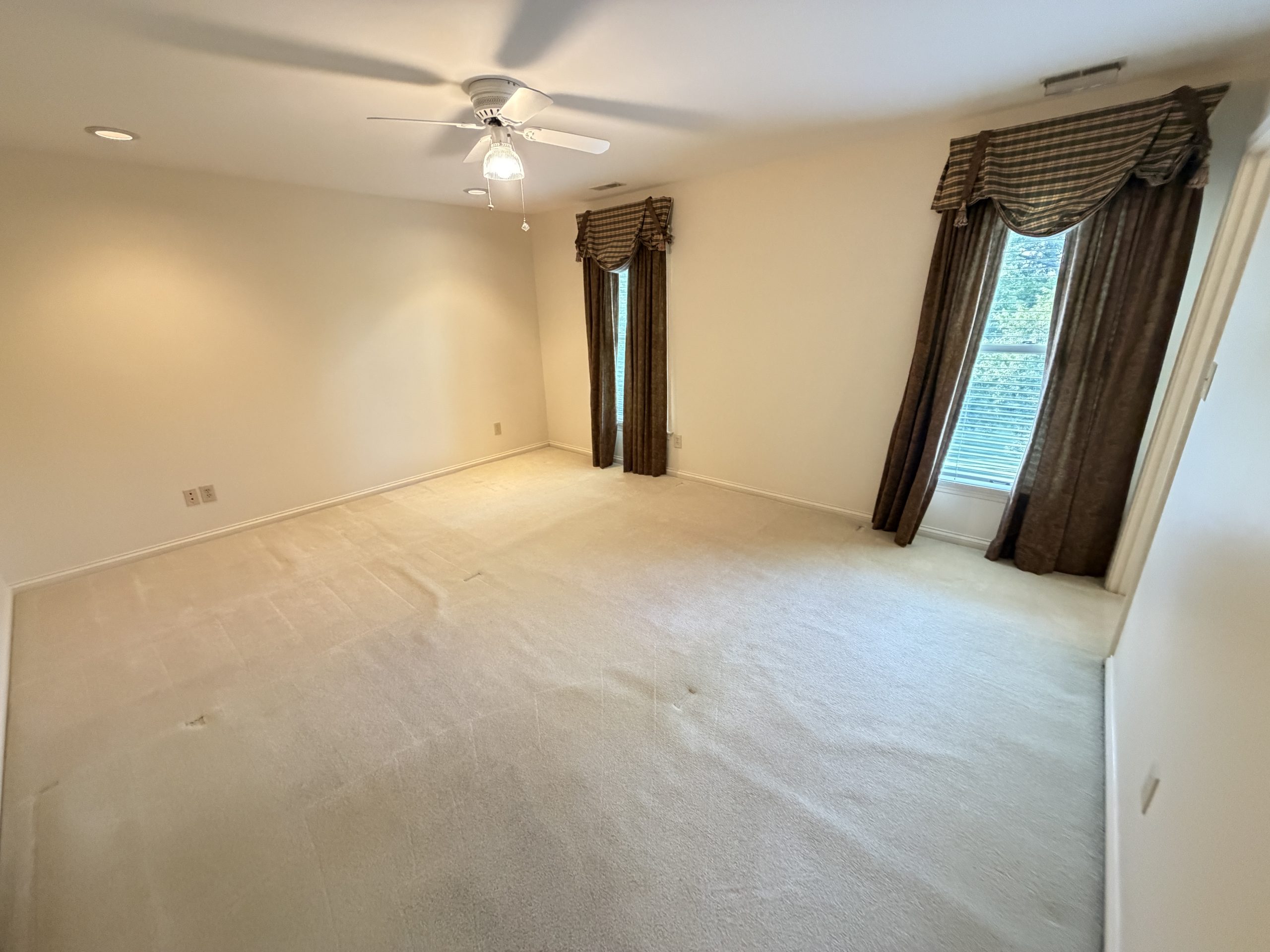 Empty beige bedroom with a ceiling fan and two tall windows covered by brown drapes.