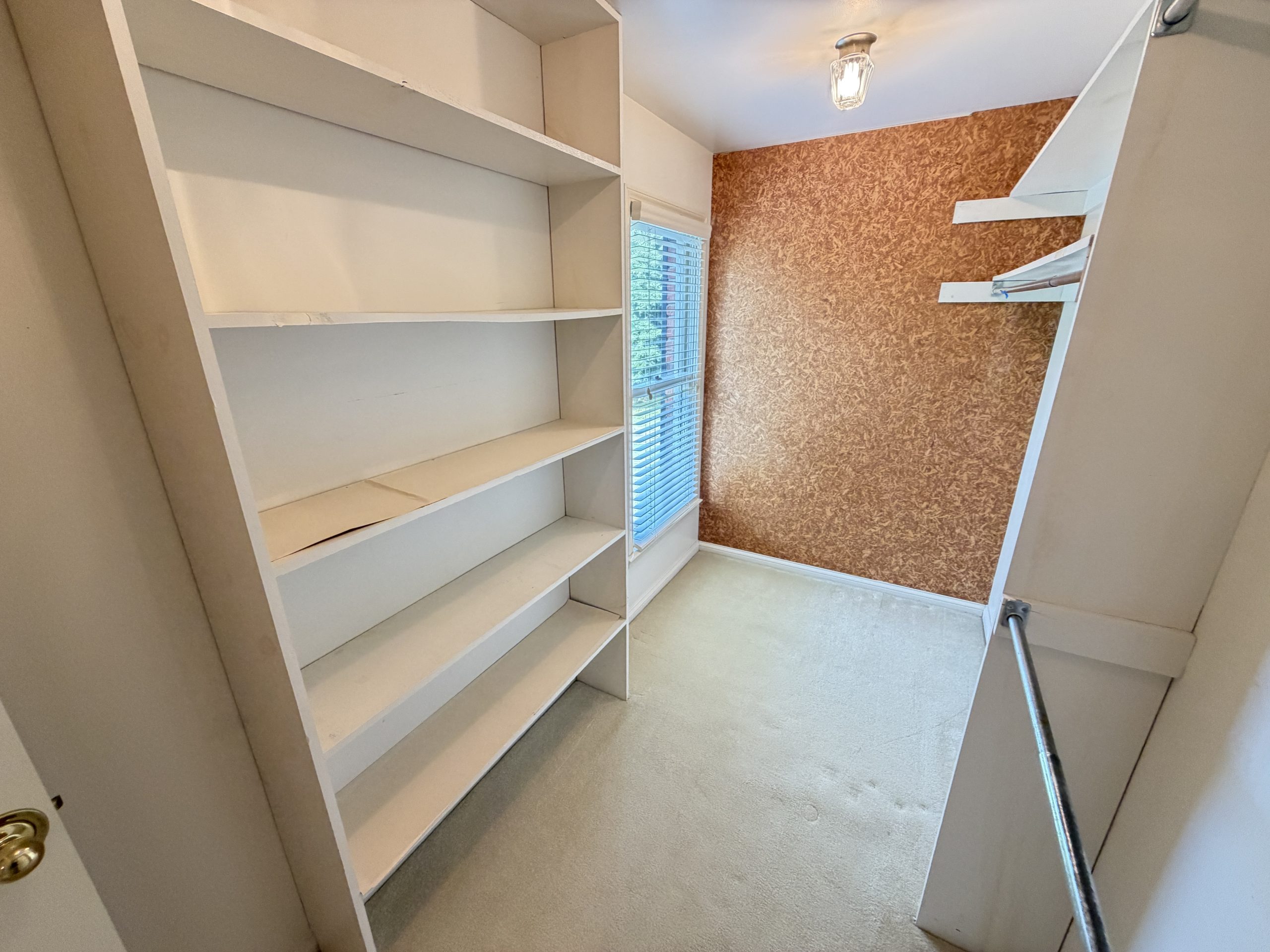 Narrow pantry with white built-in shelves on the left, a window with blinds, and brown patterned wallpaper on the right wall; a metal handrail along the right edge.