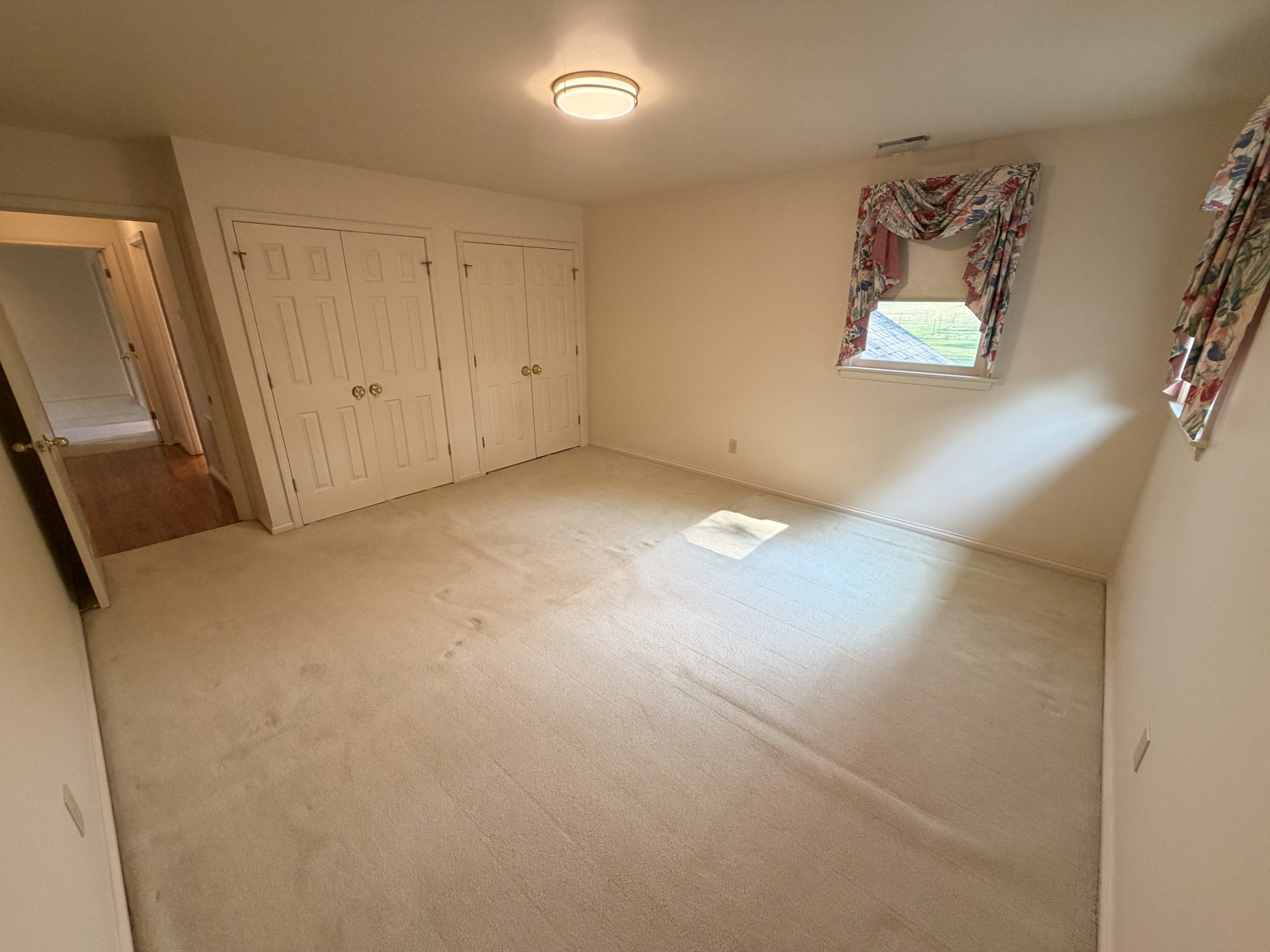Beige carpeted bedroom with two white closet doors, a round ceiling light, and a small window with floral curtains showing a yard outside.