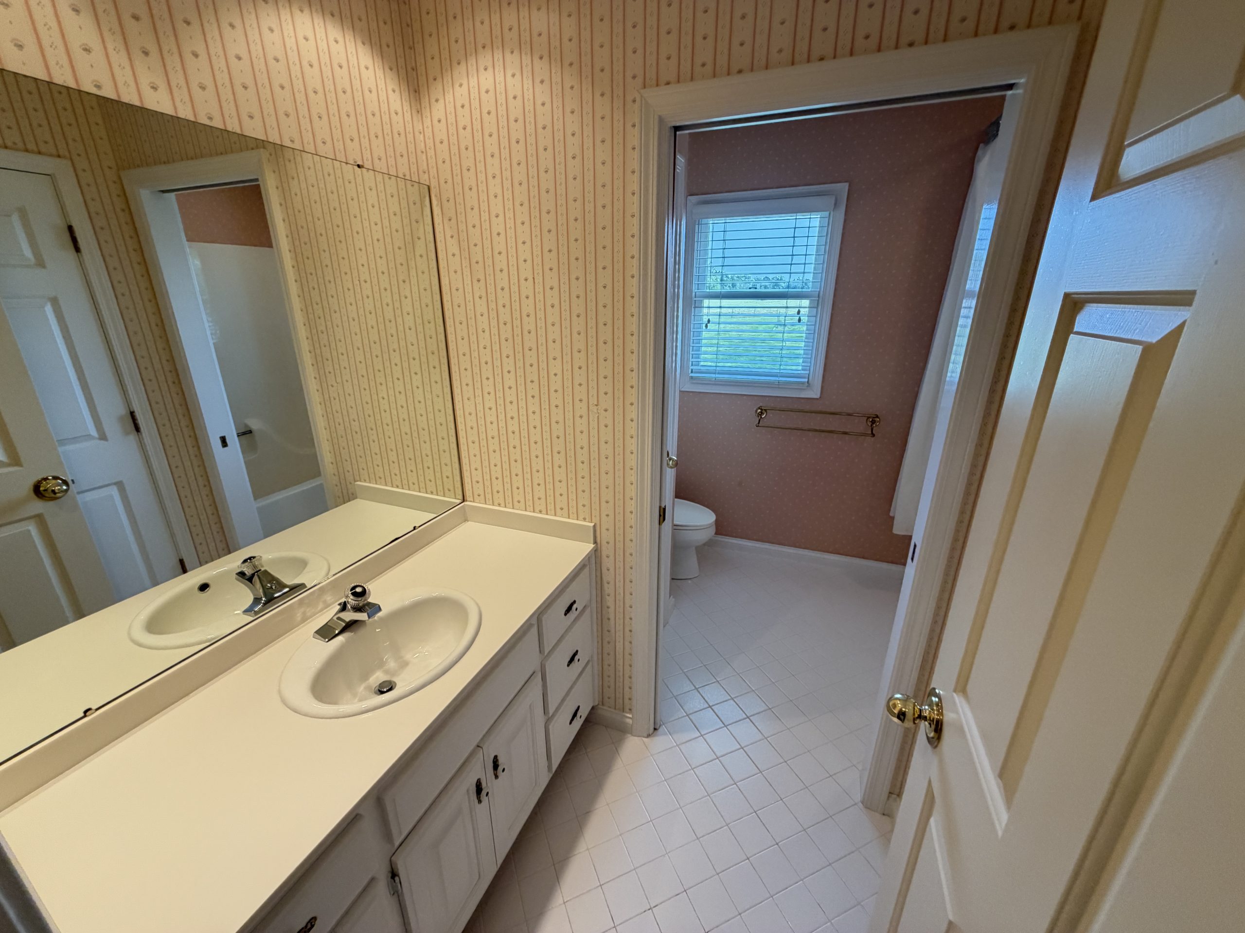 Bathroom with a white double vanity and dual sinks, large mirror, and pink wallpaper; doorway leads to a toilet and windowed room.