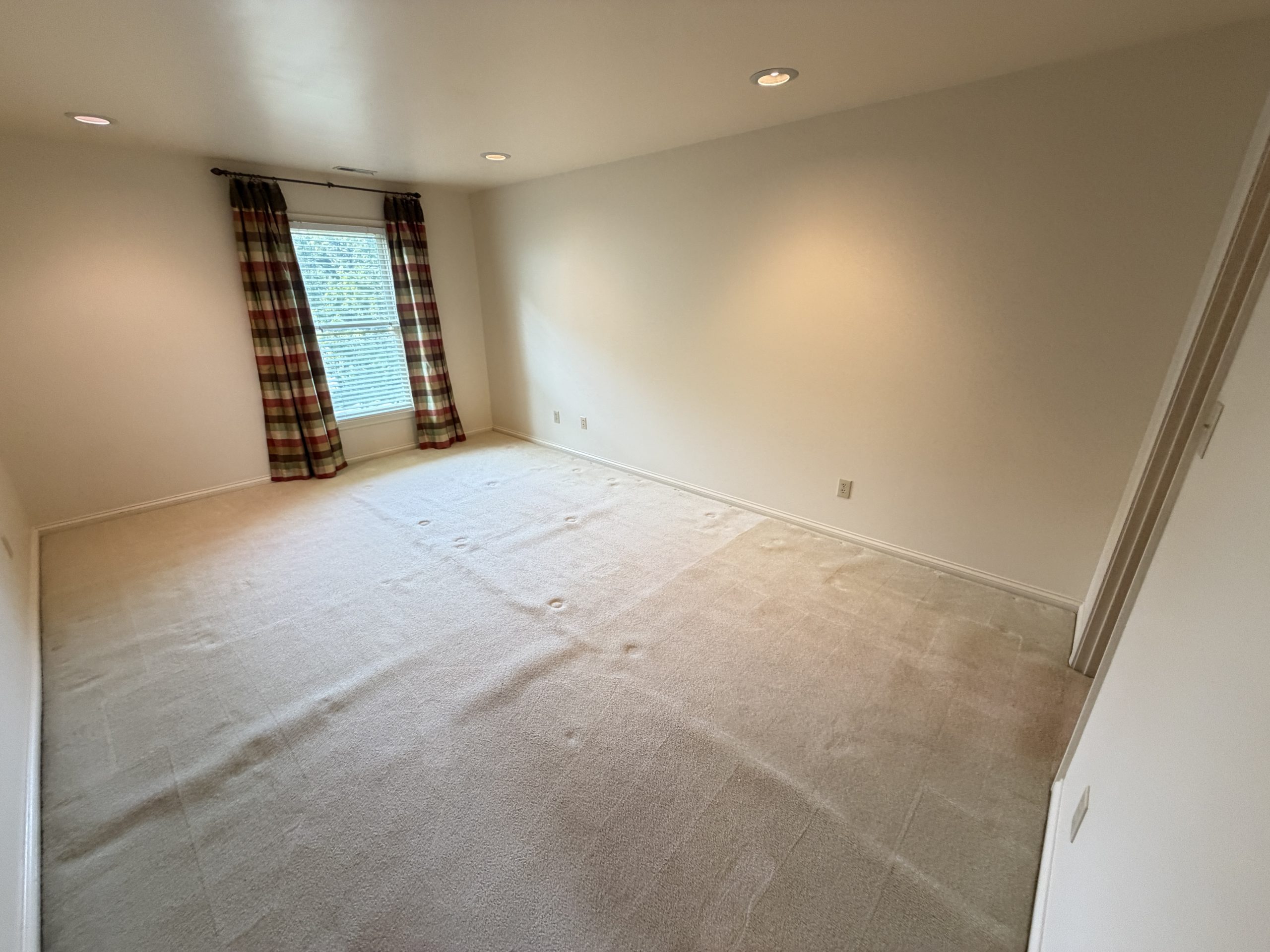 Empty beige carpeted room with a window and plaid curtains; recessed ceiling lights and light-colored walls visible. (informative)