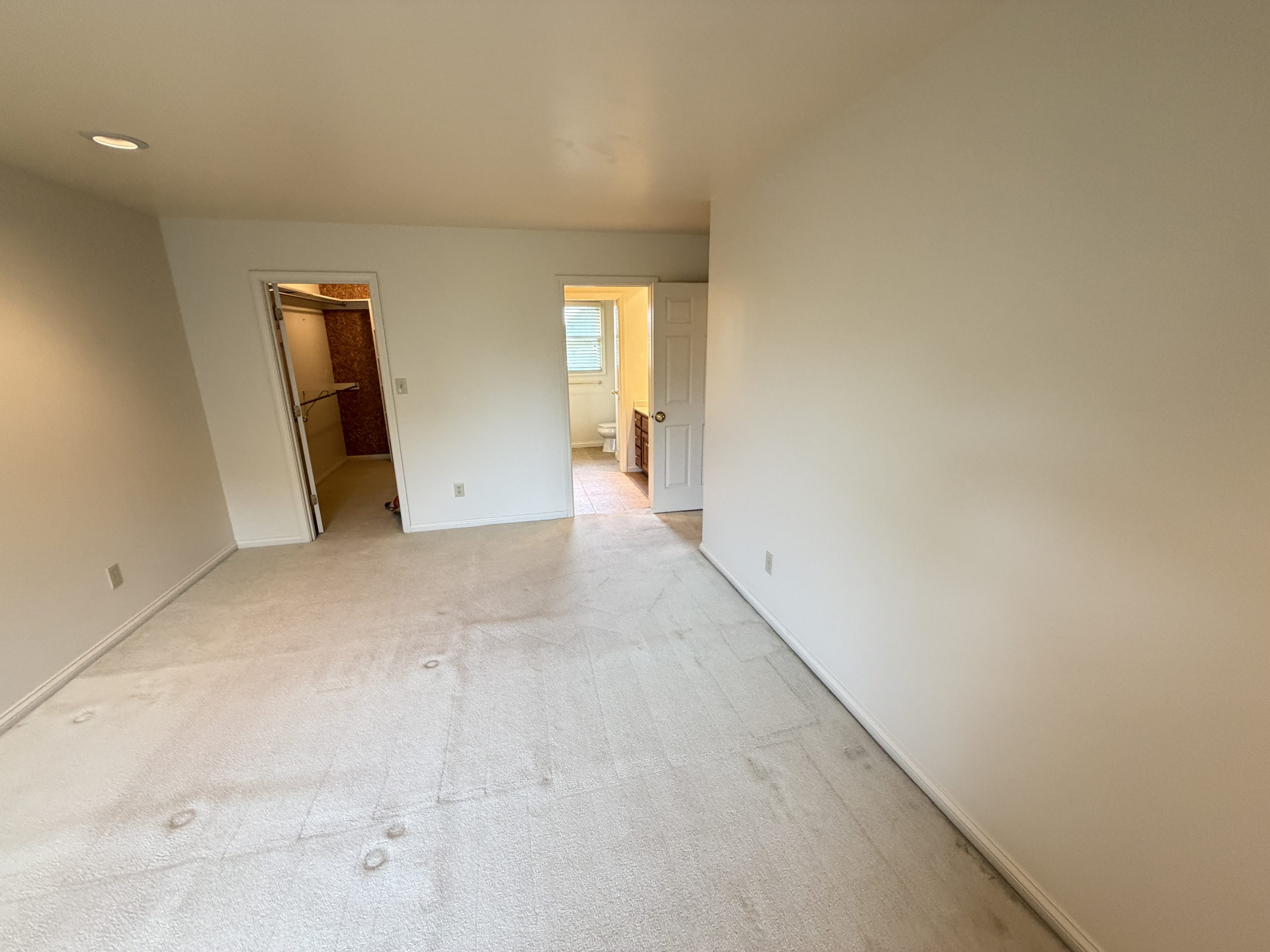 Empty beige-carpeted room with two open doors revealing a bathroom and a closet; walls are bare and a small vent is in the ceiling.