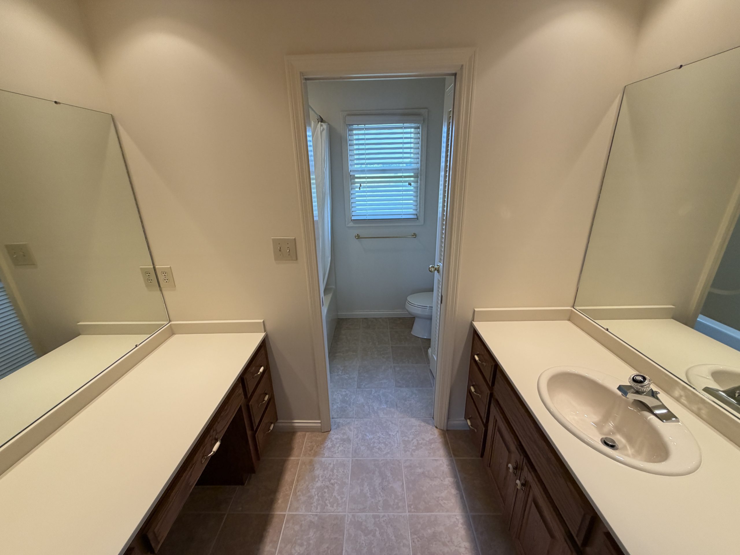 Symmetrical bathroom with two sinks and dark wooden cabinets, beige countertops, and large mirrors, doorway to a toilet at the back.