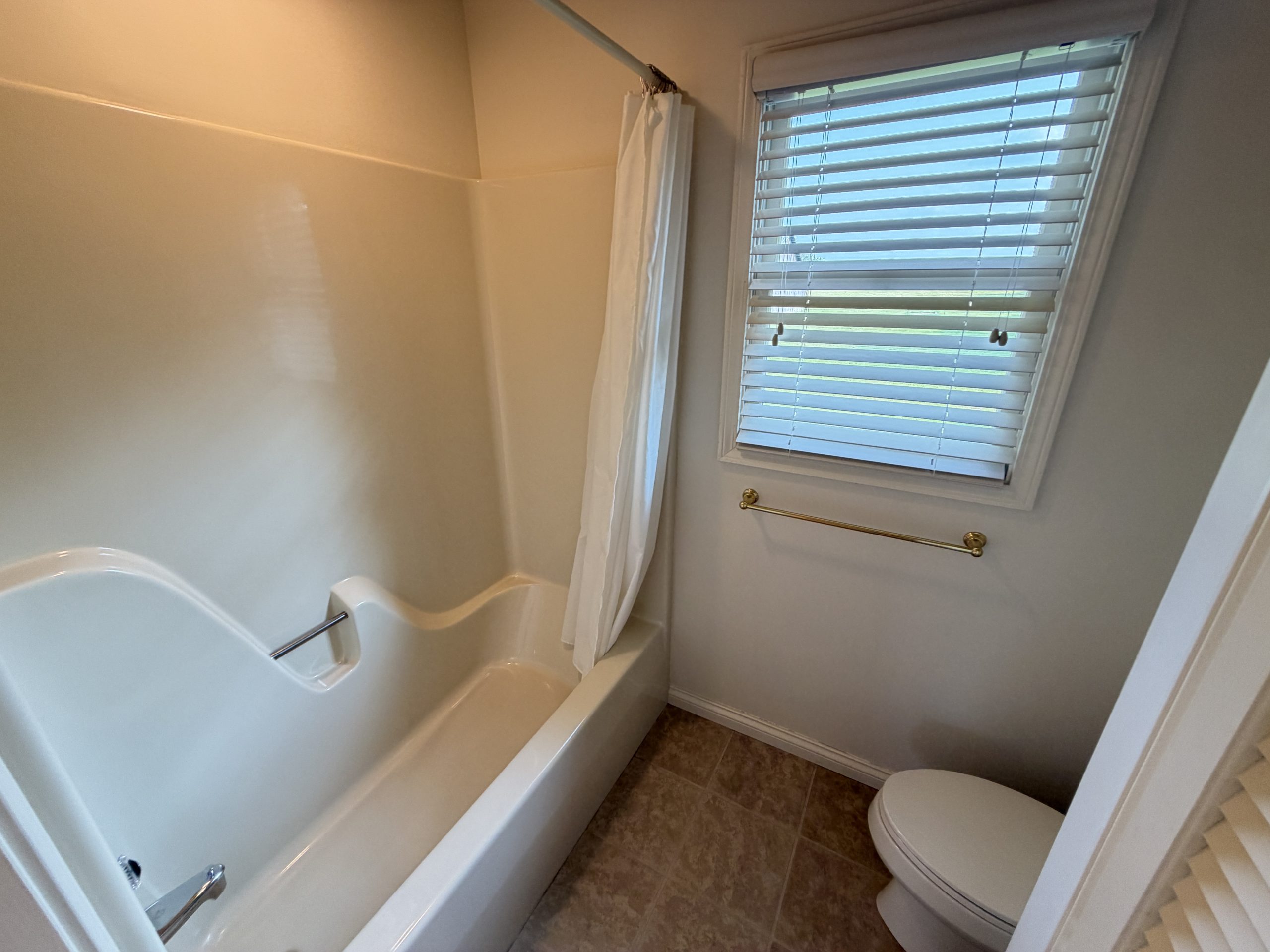 White bathtub with a curved edge and a white shower curtain next to a window with horizontal blinds in a small bathroom view from above the tub edge.