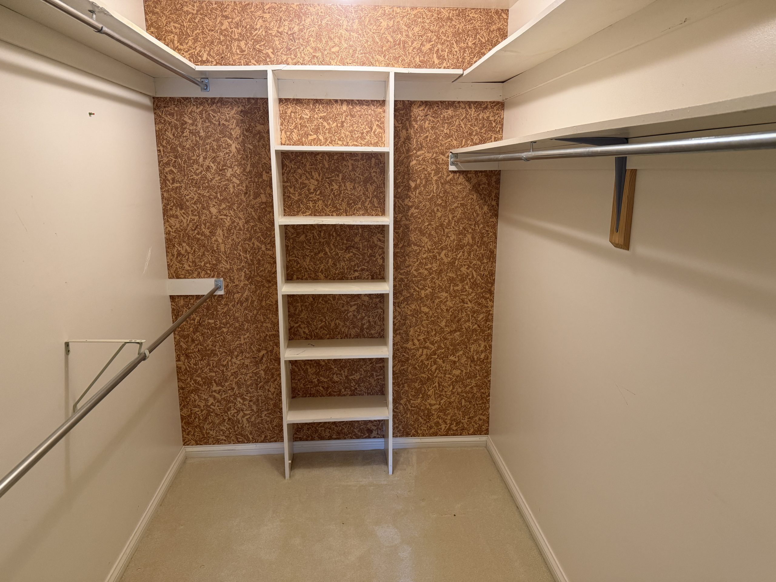Narrow walk-in closet with a white ladder-style shelf centered between brown patterned wallpaper and metal clothing rods on both sides.