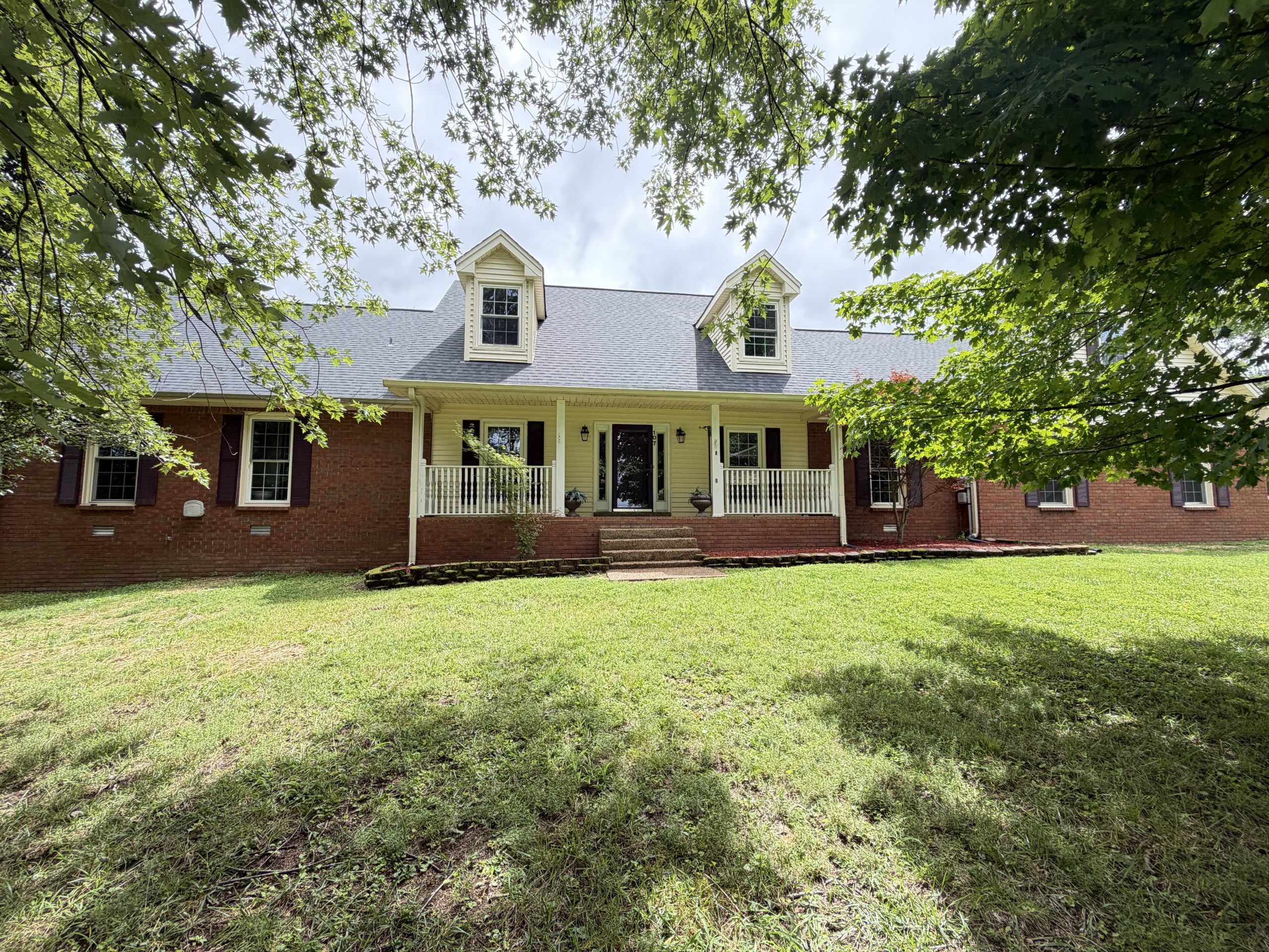 Front view of a brick-and-siding house with dormer windows, a covered porch, and a lawn framed by leafy trees